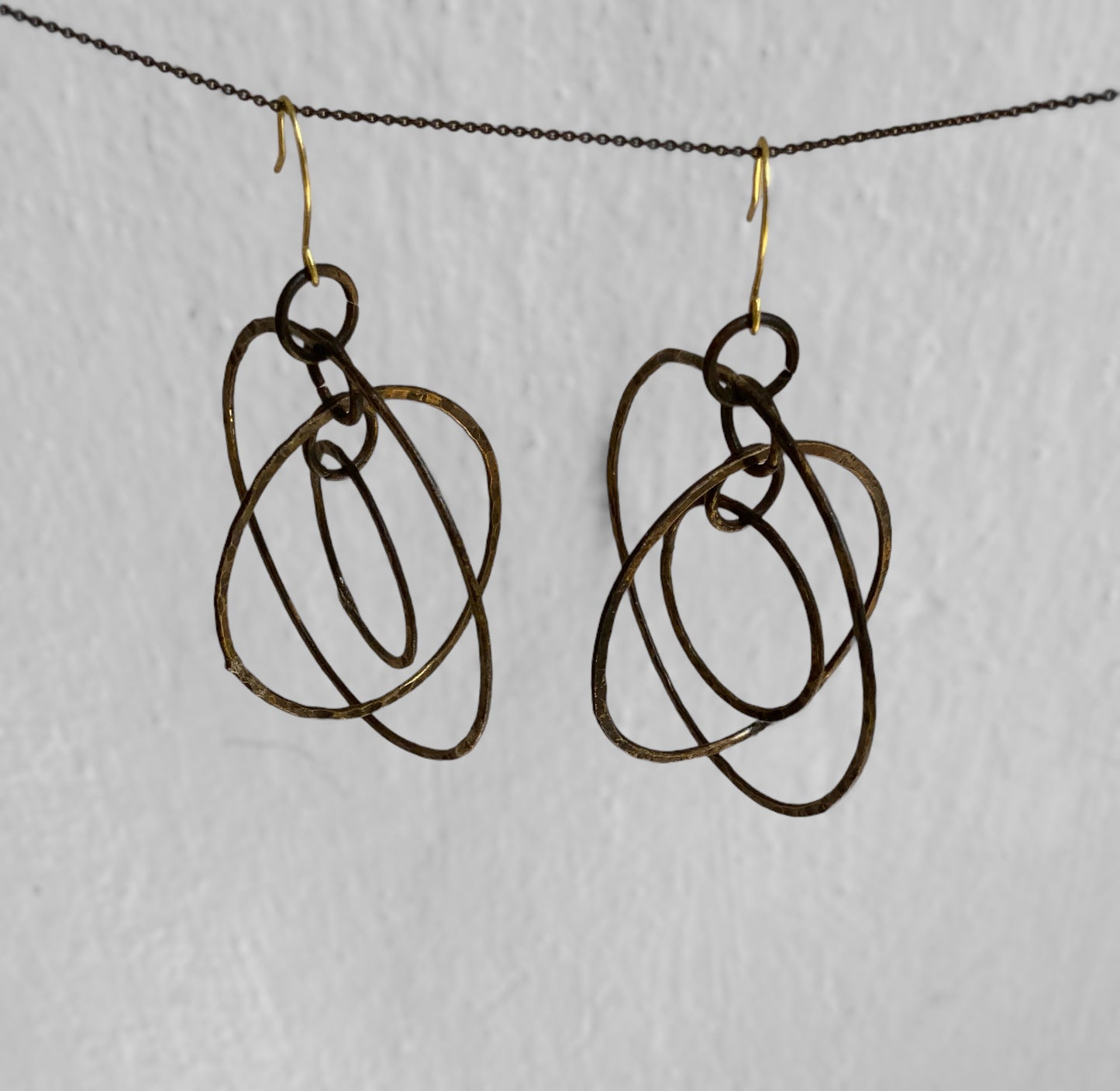 A pair of handcrafted earrings featuring abstract hammered brass loops, suspended on gold hooks, styled on a neutral background to highlight their artistic and unconventional design.