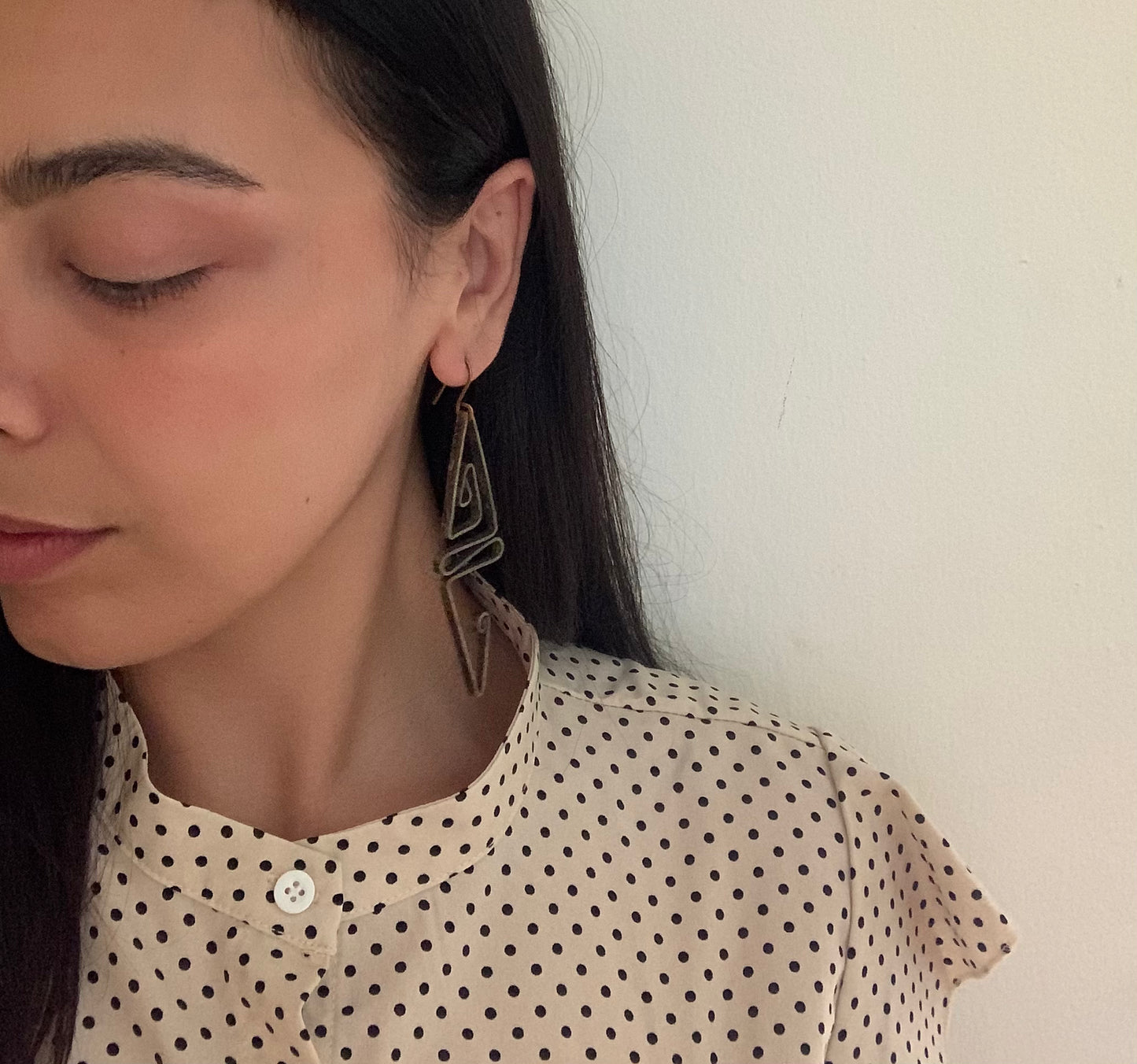 A close-up of a model wearing handcrafted abstract brass structural earrings with a geometric design, styled with a minimalist polka-dot blouse for a modern and elegant look.