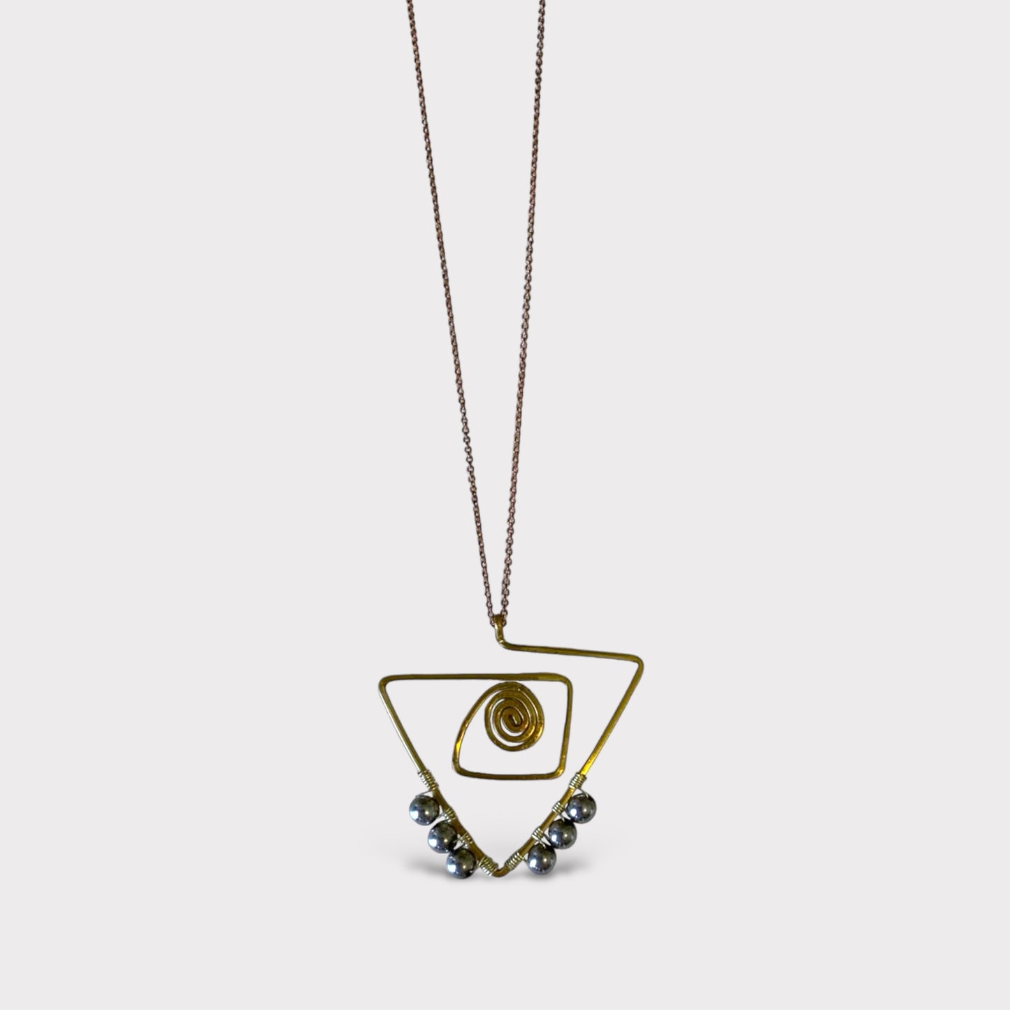 A handcrafted brass necklace featuring an abstract triangular design with a spiral centerpiece and metallic bead accents, suspended on a delicate chain, displayed on a clean white background