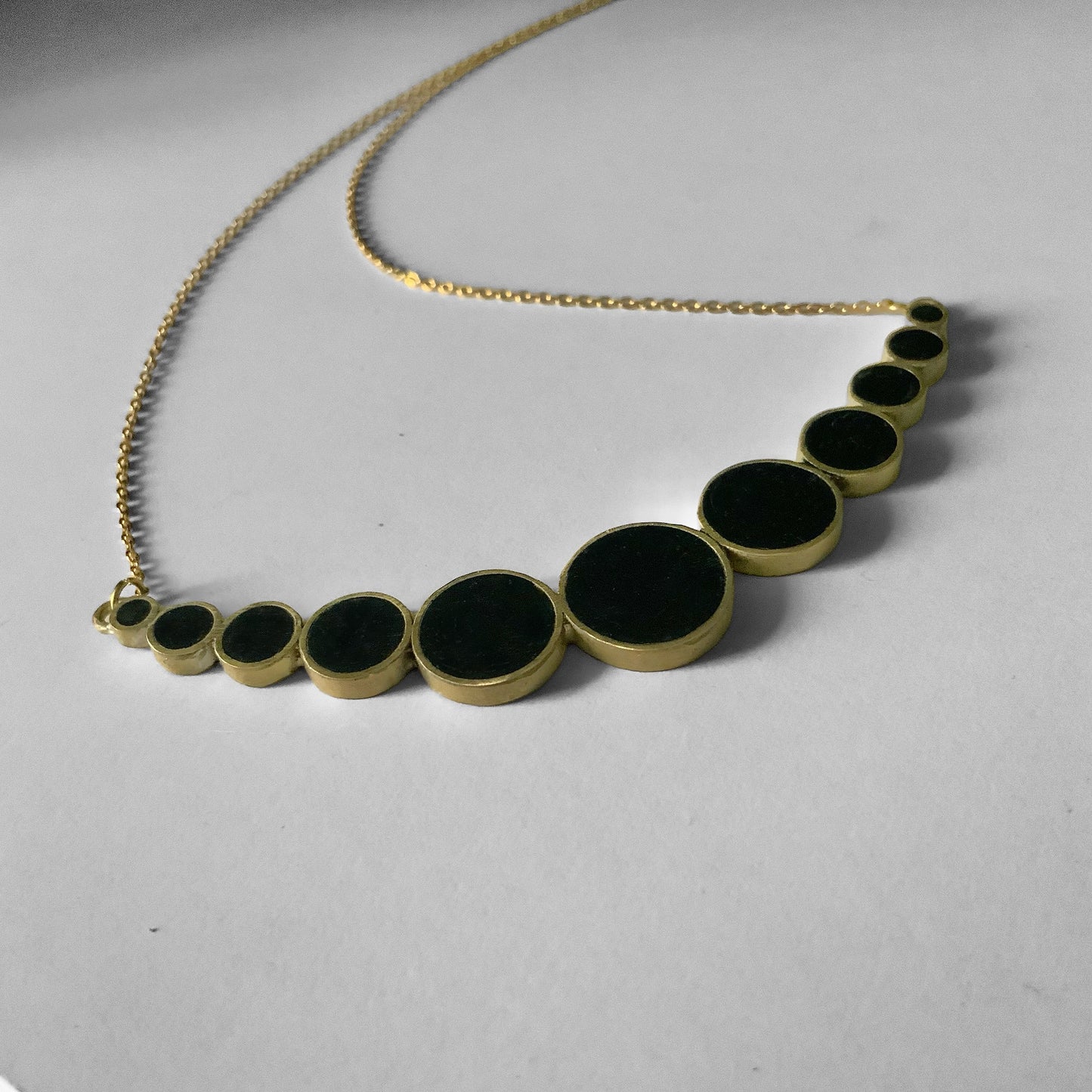 A handcrafted brass necklace featuring a gradient design of black circular accents in varying sizes, arranged in a semi-curve, suspended on a delicate staiknless steel chain, displayed on a light background.