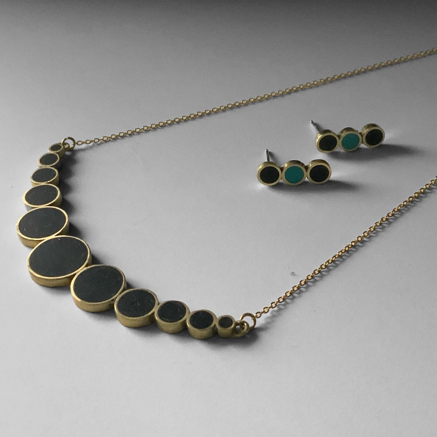 A brass necklace featuring black circular accents in a gradient design, paired with matching earrings, displayed on a light surface for a cohesive set presentation.