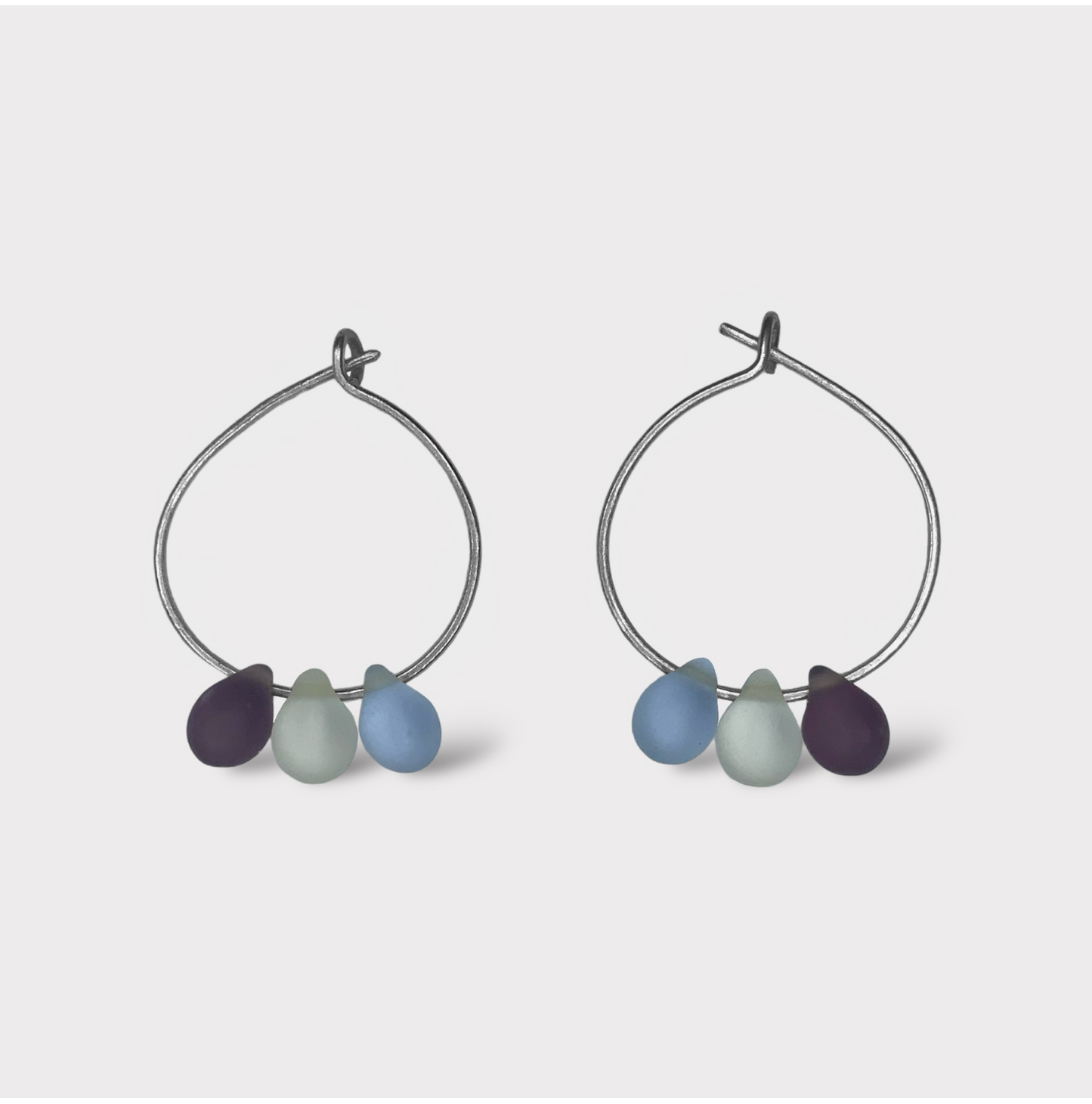 A pair of silver hoop earrings adorned with three seaglass pendants in shades of deep blue, light blue, and purple, inspired by the refreshing hues of Greek summer.