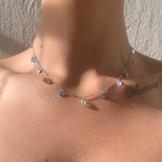 A dainty silver chain necklace adorned with small, colorful seaglass charms in shades of green, blue, and white, styled on a model's collarbone and illuminated by soft natural sunlight. A minimalist and elegant design inspired by the vibrant hues of a Greek summer.