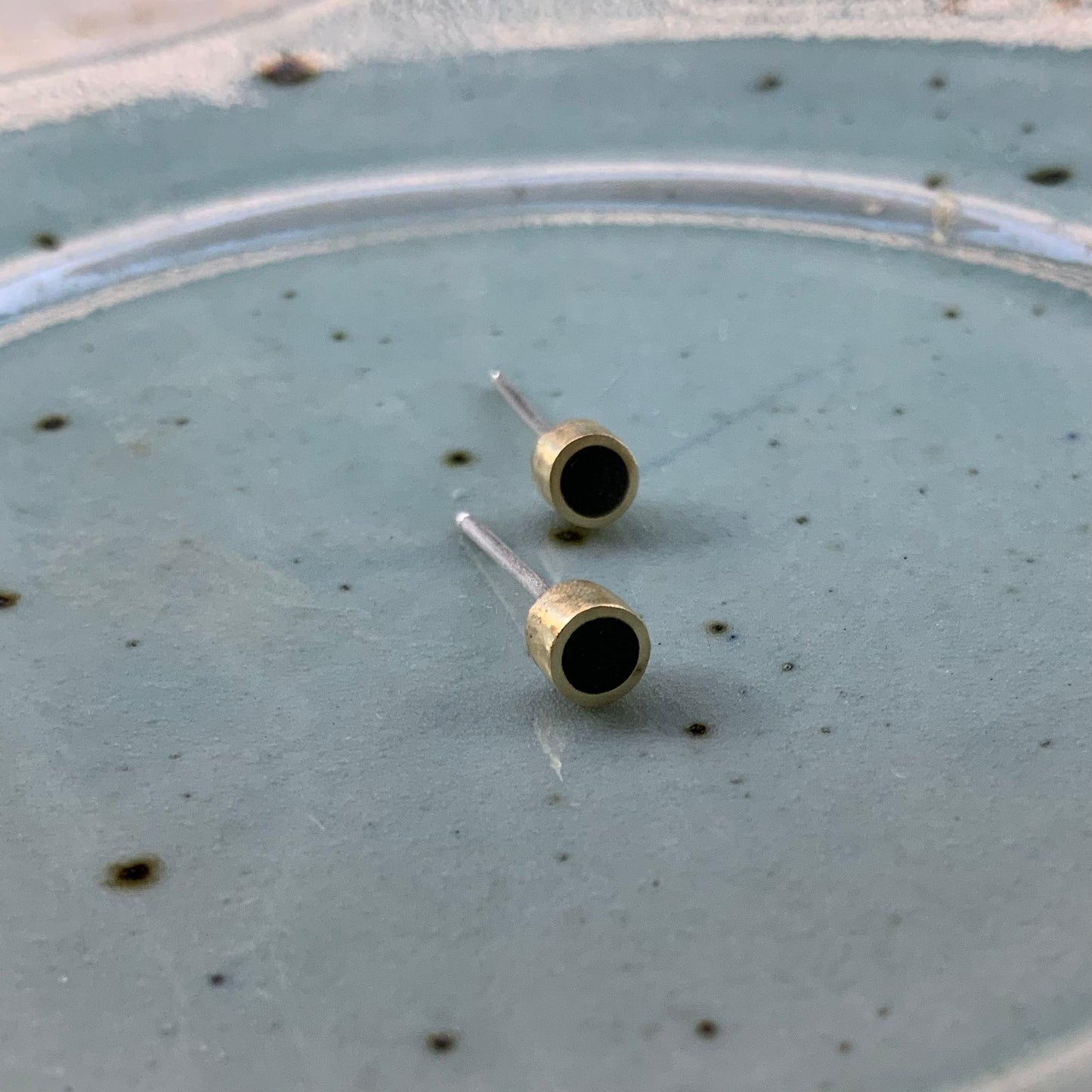 A pair of brass stud earrings with black circular inlays, elegantly presented on a speckled ceramic plate for a rustic and artistic touch.