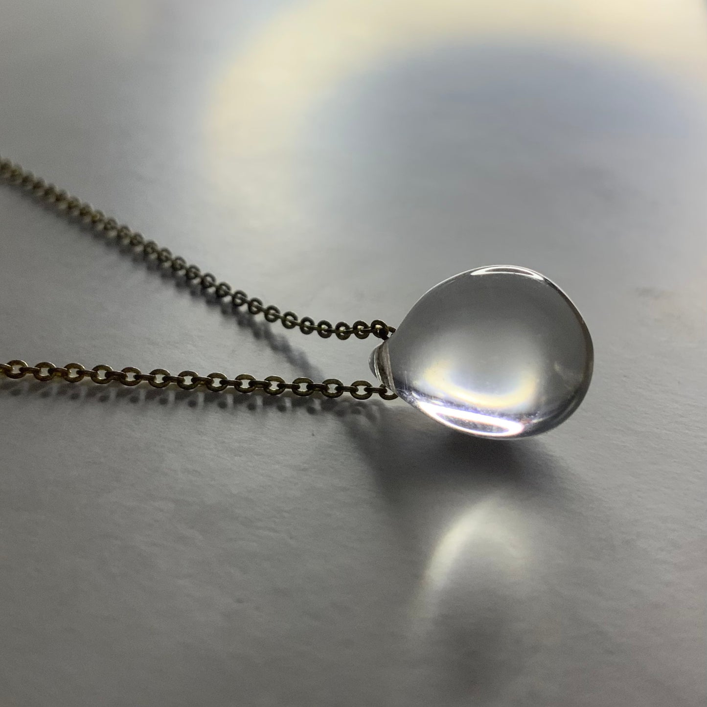 A minimalist necklace featuring a clear translucent teardrop glass pendant, elegantly suspended on a brass chain, styled on a softly lit surface for a modern and elegant look.