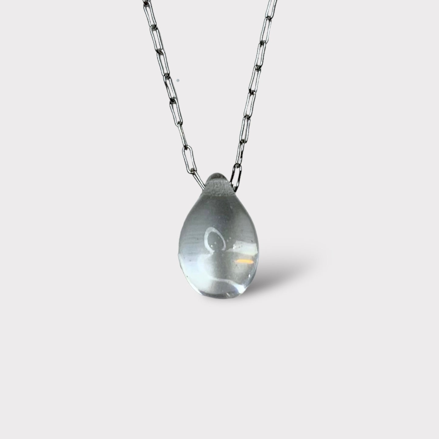 A minimalist necklace featuring a clear translucent teardrop-shaped glass pendant, elegantly suspended on a stainless steel chain, styled on a white background.