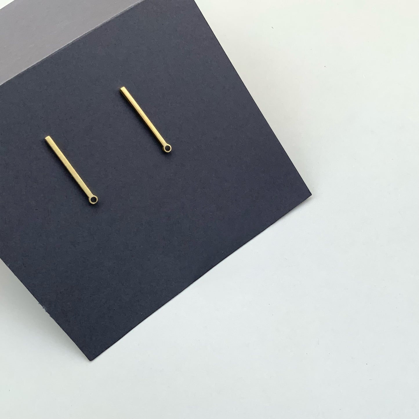 A close-up of brass bar earrings with a circular cutout at the base, photographed on a black stand againt a white background for a clean and elegant look