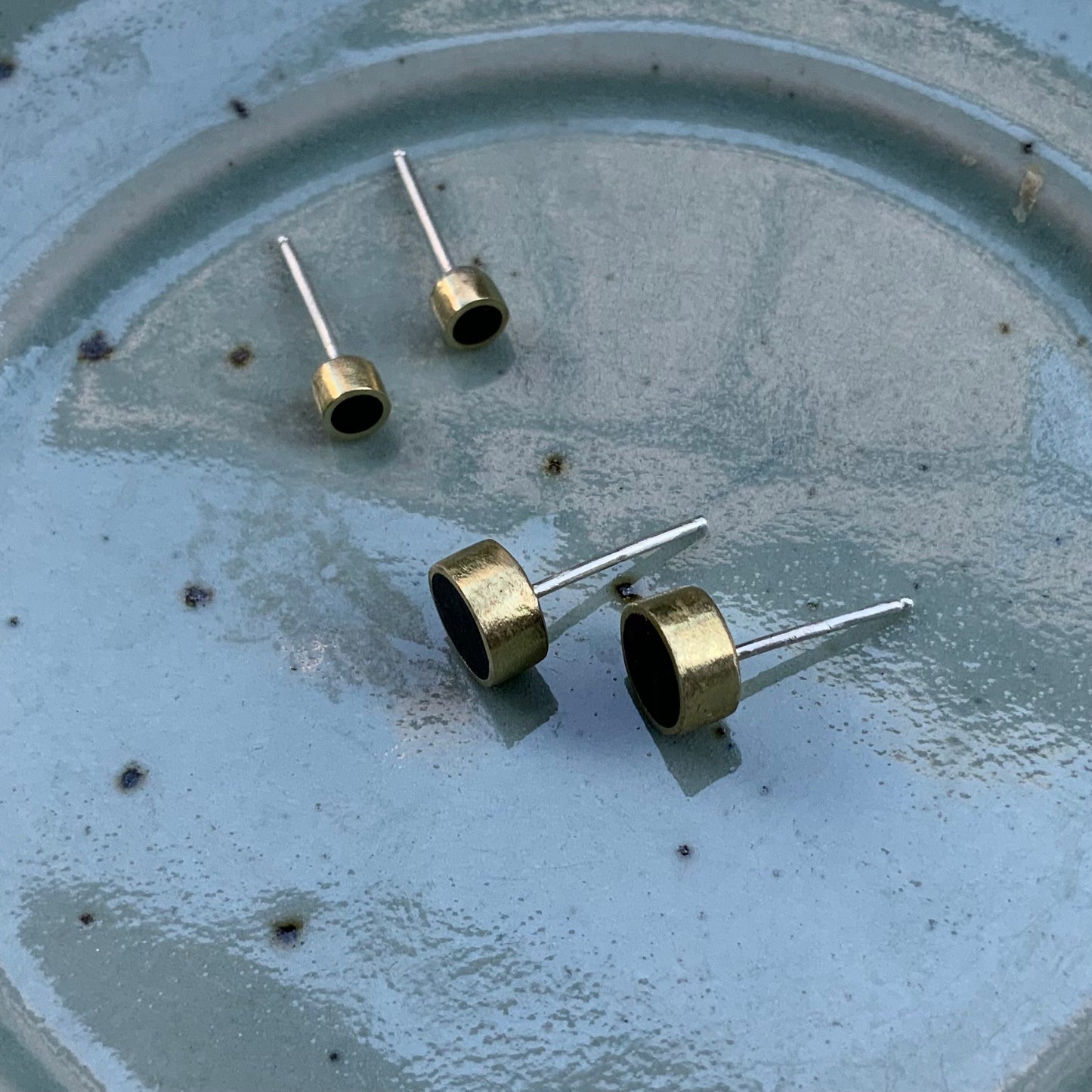 Two pairs of handcrafted brass stud earrings featuring black circular inlays, elegantly arranged on a speckled ceramic plate, showcasing their minimalist and geometric design.
