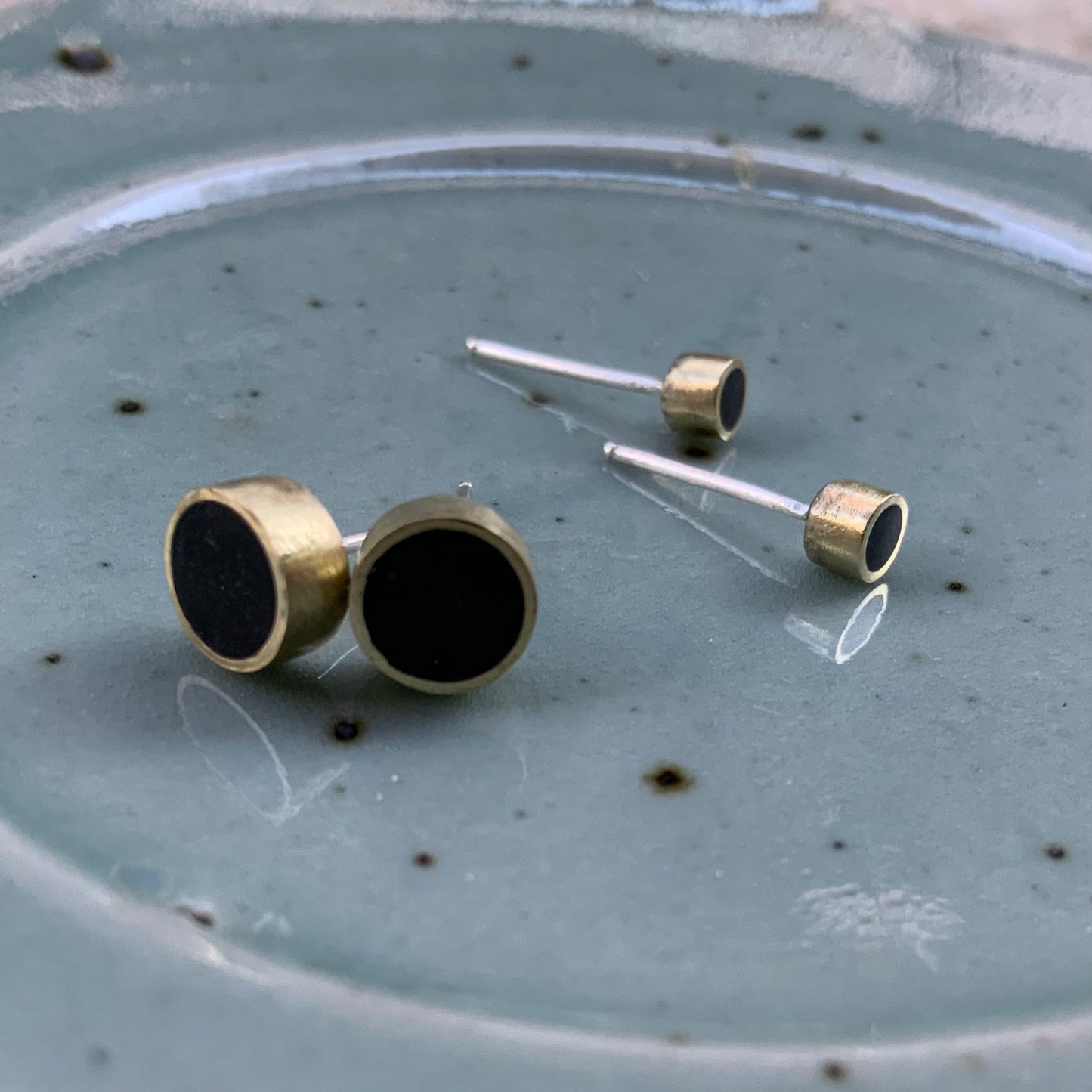 Two pairs of brass stud earrings with silver posts and black circular polymer clay inlays, elegantly arranged on a speckled ceramic plate for a rustic and artistic presentation