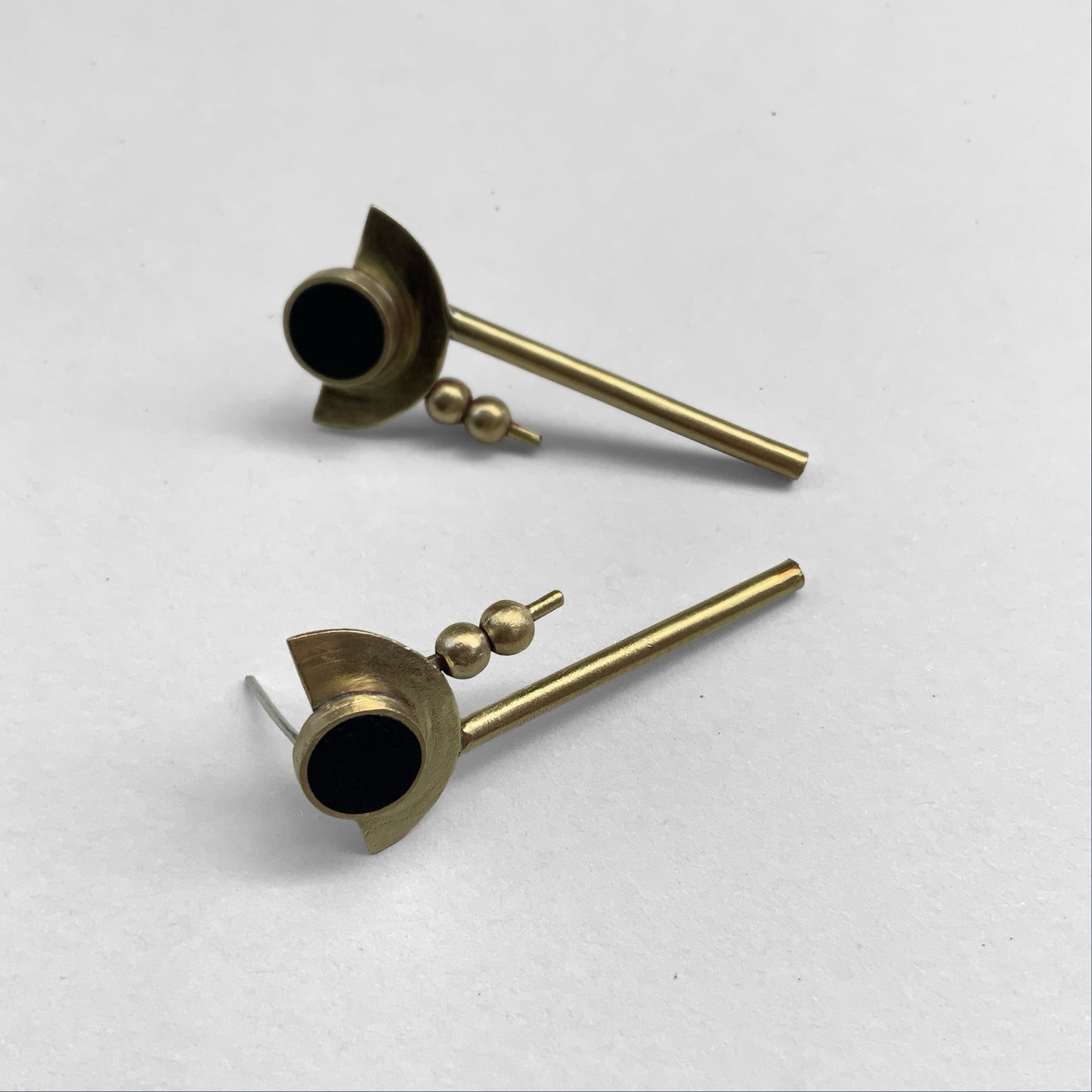 Geometric drop earrings with a black circular centerpiece framed by crescent brass details, two small brass beads, and a vertical brass rod, elegantly arranged on a white surface