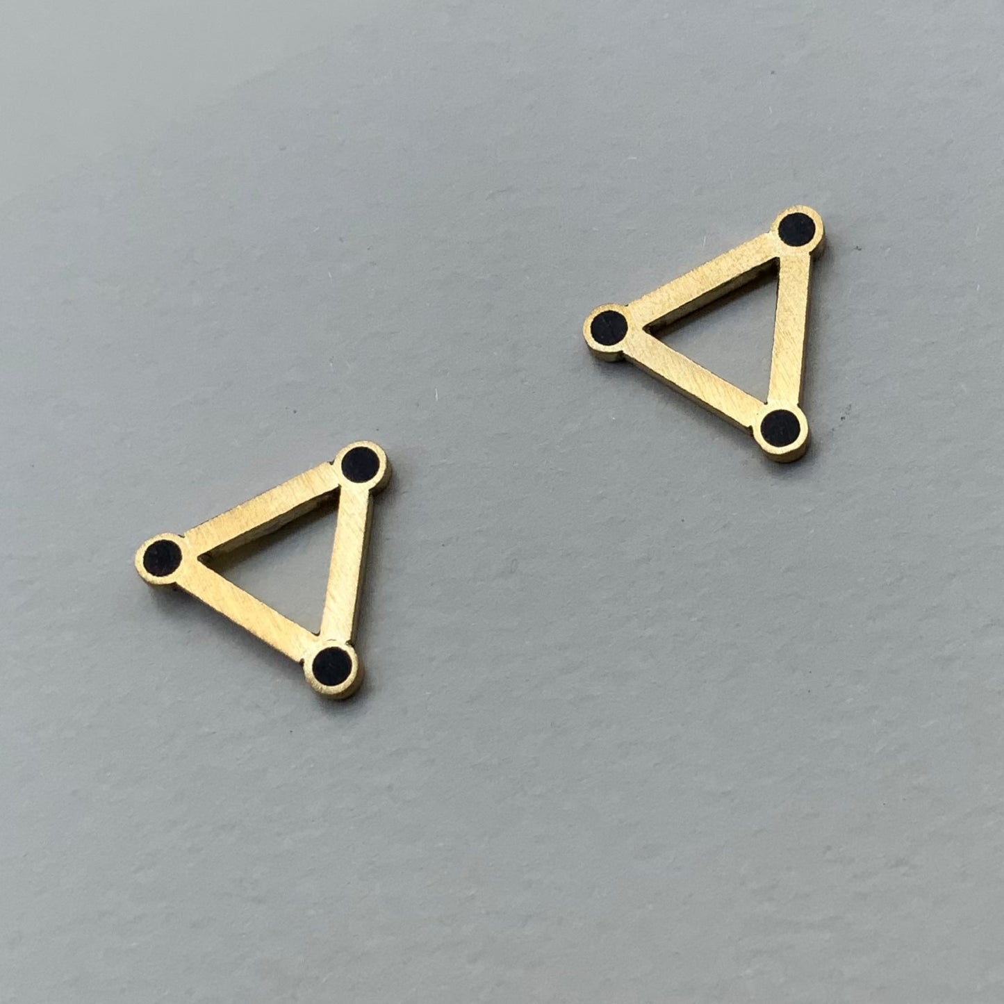 A pair of handcrafted brass triangle earrings with black circular accents at each corner, displayed on a light gray background to highlight their modern and geometric design