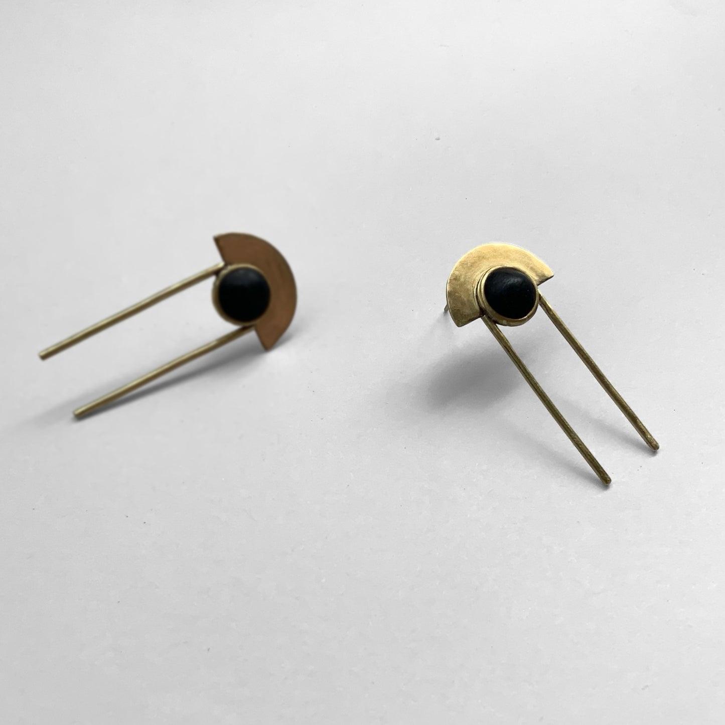 A pair of geometric earrings with black circular accents and brass details, complemented by sleek, minimalist rod drops, photographed on a light gray background