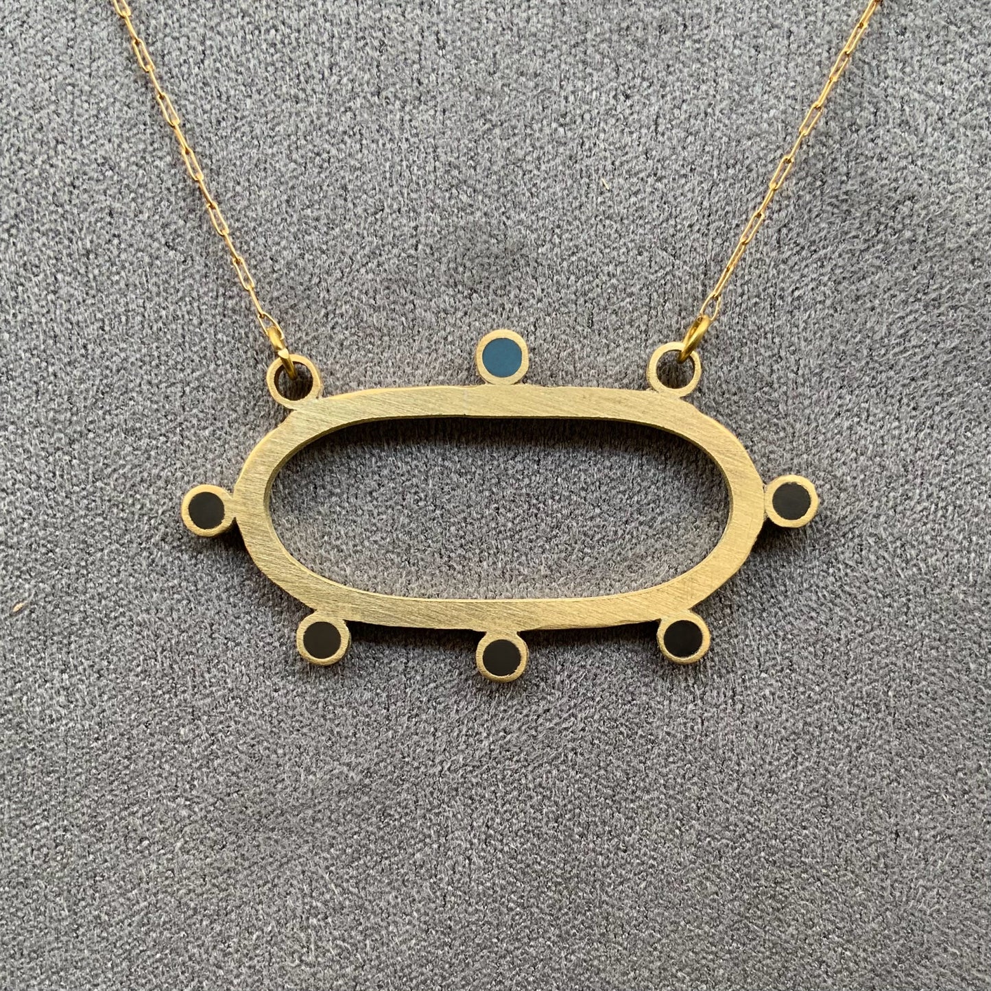 A modern brass necklace showcasing an oval-shaped pendant adorned with circular cutouts and a single blue accent, hanging from a fine stainless steel chain, styled on a gray fabric surface.