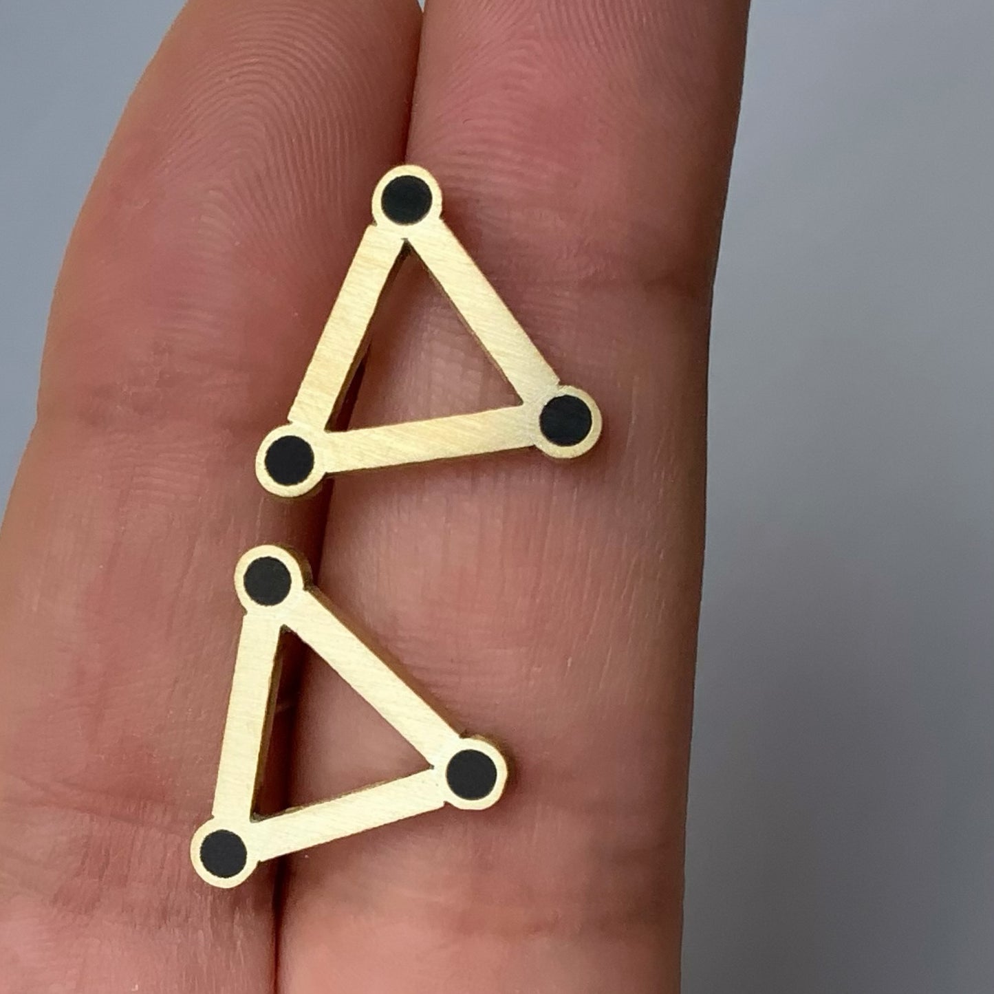 Handcrafted brass triangle earrings featuring black circular accents, held between fingers to showcase their size and lightweight design.