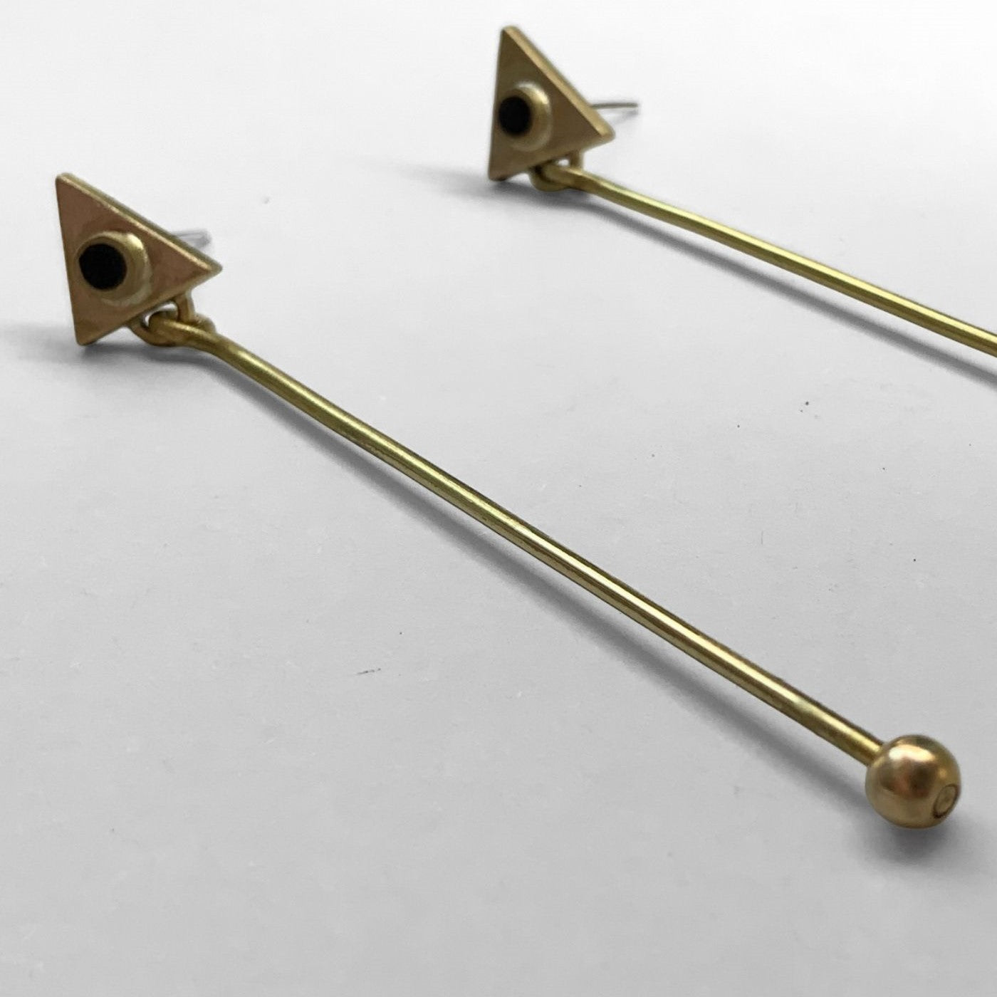 A close-up of geometric brass earrings with a triangle stud, black circular accents, and elongated brass rods with ball tips, photographed on a light gray surface