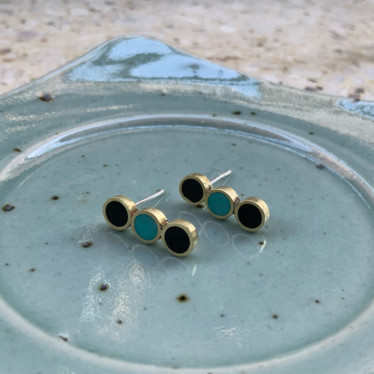 Brass stud earrings with three circular accents—two black and one turquoise—elegantly arranged on a speckled ceramic plate, showcasing their minimalist and modern design