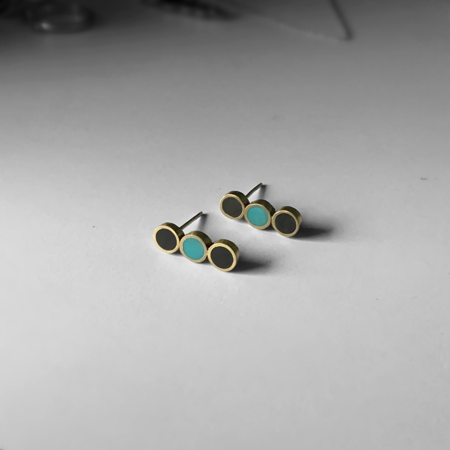 A pair of brass stud earrings with three circular accents—black and turquoise—arranged in a minimalist linear design, displayed on a light gray surface with subtle background details
