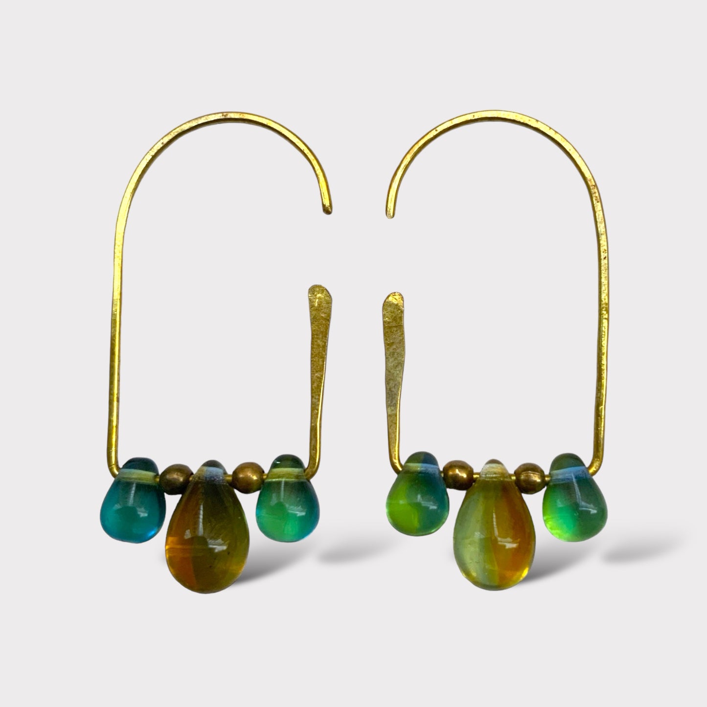 A pair of golden handcrafted hoop earrings featuring gradient teardrop-shaped glass pendants in shades of green, blue, and amber, accented with brass beads, displayed on a white background for a minimalist look.