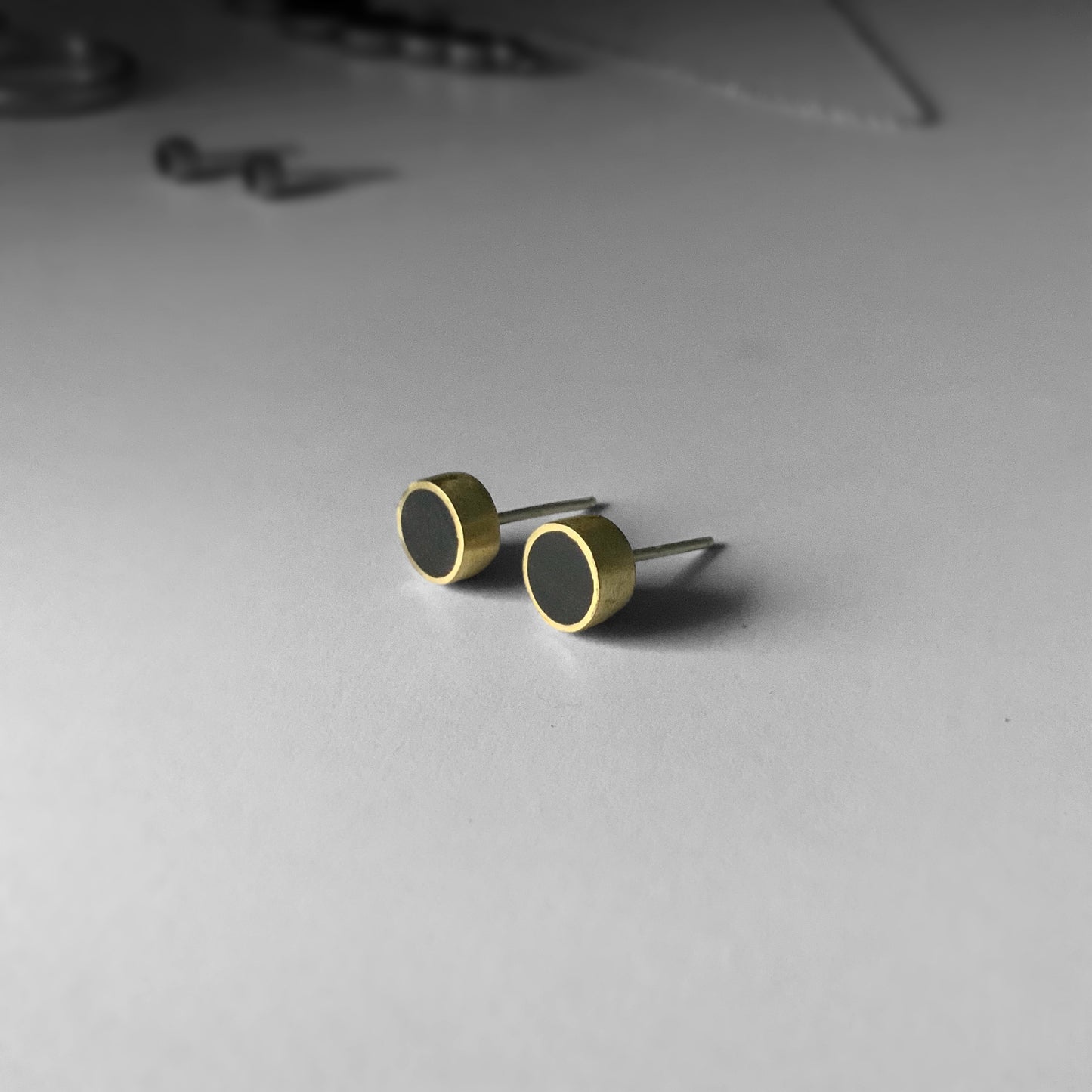 A pair of handcrafted brass stud earrings featuring black circular accents, styled on a light gray surface with subtle background details to highlight their minimalist aesthetic.