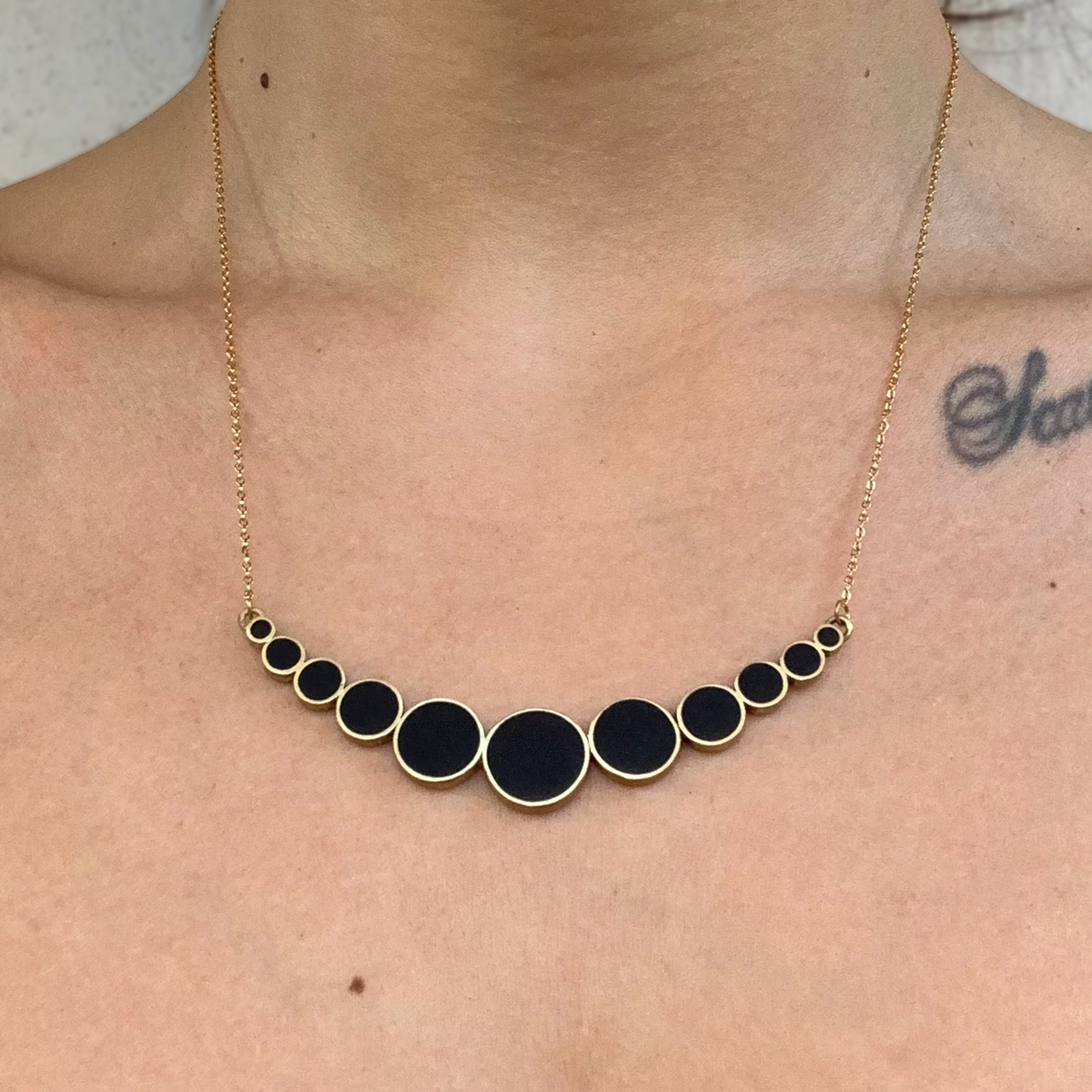A brass necklace with black circular accents in a gradient design, worn by a model to showcase its fit and elegant curve along the neckline.