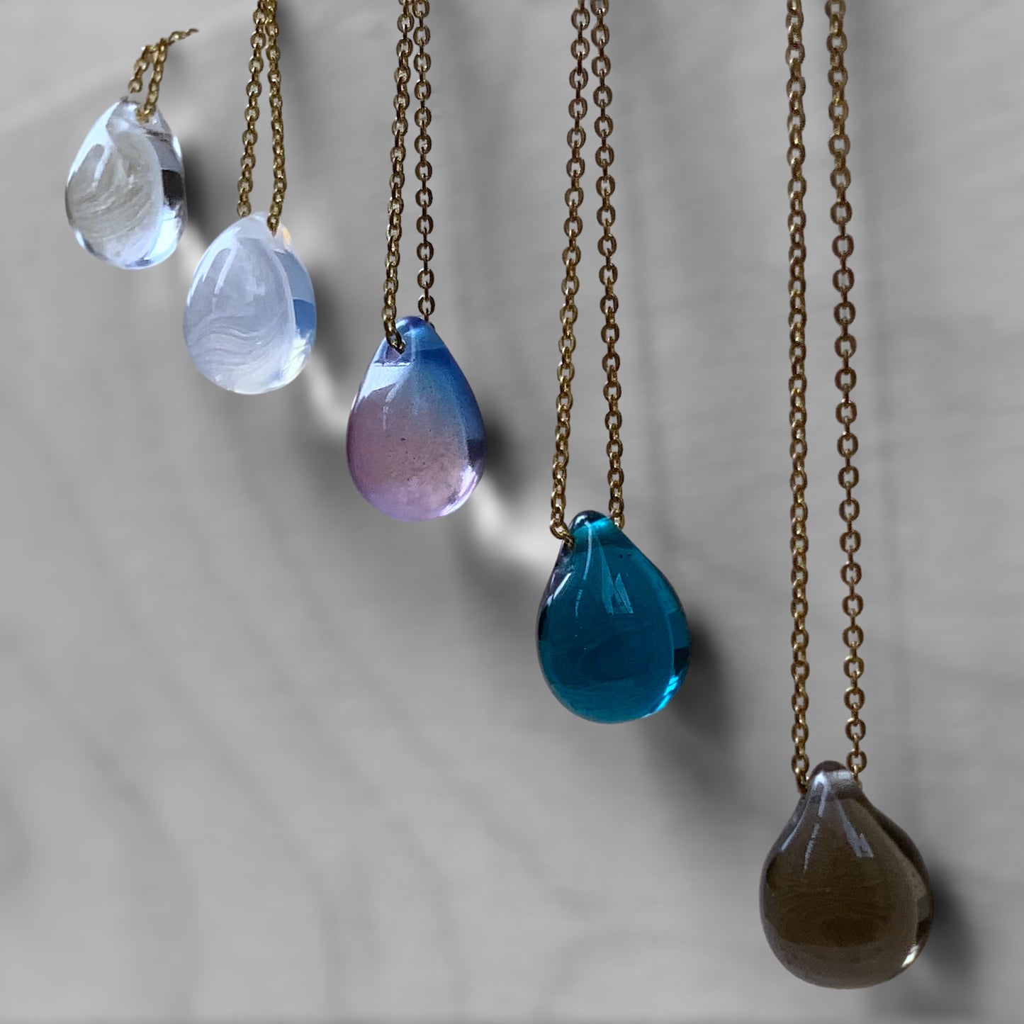 A collection of handcrafted necklaces featuring gradient translucent teardrop glass pendants in shades of white, purple, aqua blue, and brown, suspended on brass chains and displayed on a light gray background.