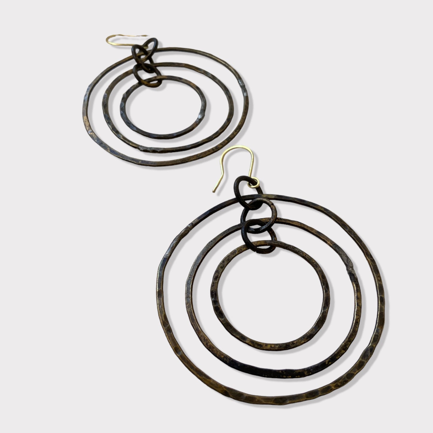 A close-up of handcrafted earrings with three concentric hammered brass circles, elegantly connected and hanging on gold hooks, displayed on a white background for a modern aesthetic.