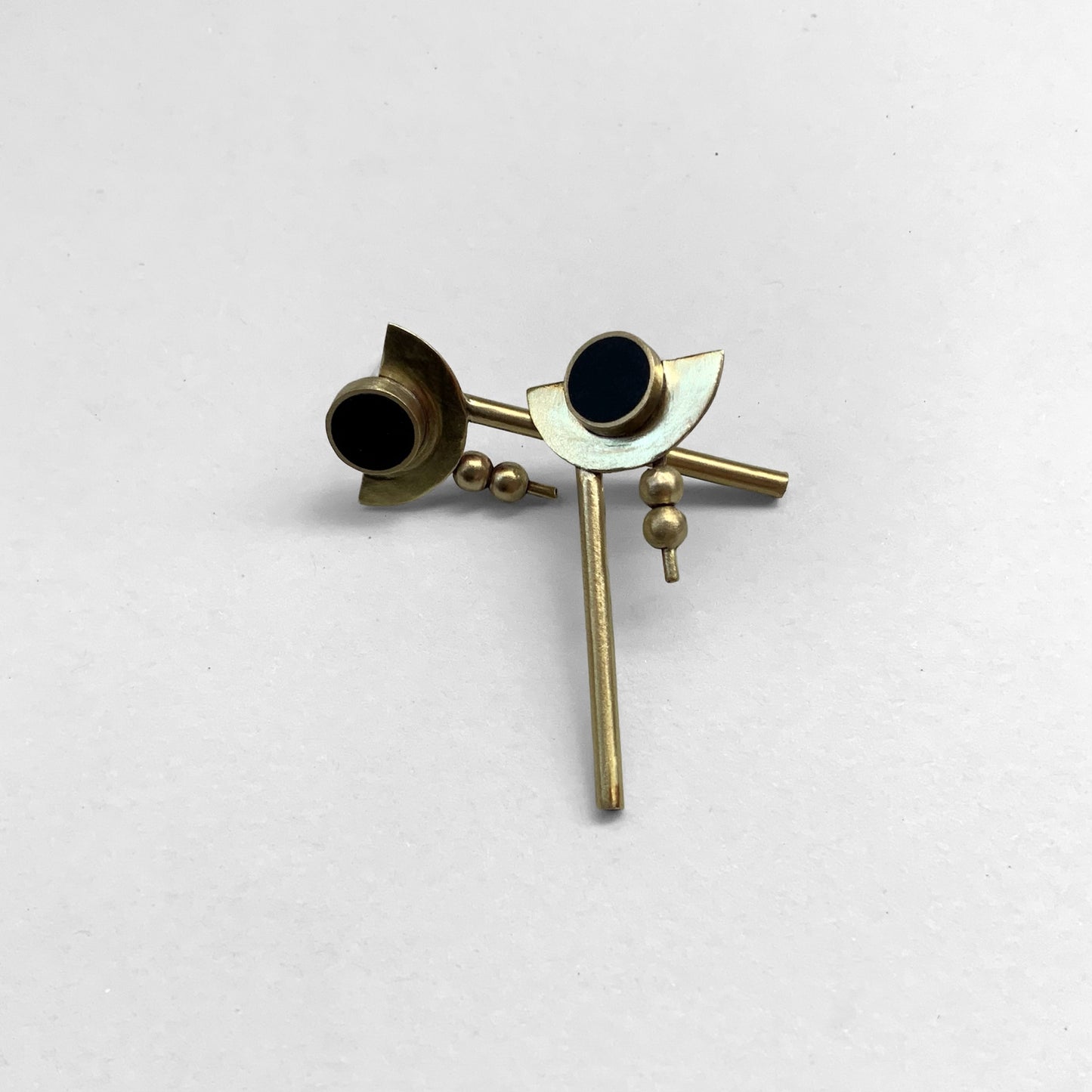 Artistic drop earrings featuring black circular accents framed by brass crescents, minimalist rods, and small brass bead details, styled in a layered arrangement on a light background