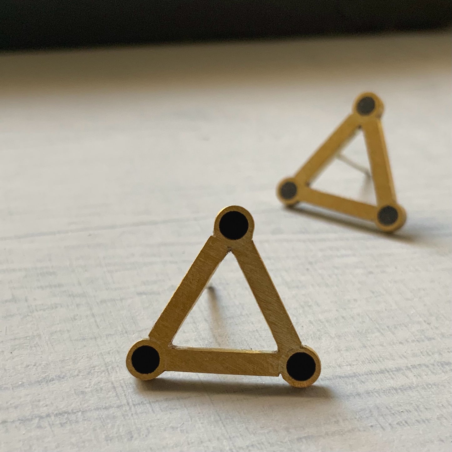 A pair of minimalist brass triangle earrings with black circular accents at each corner, displayed on a light gray background