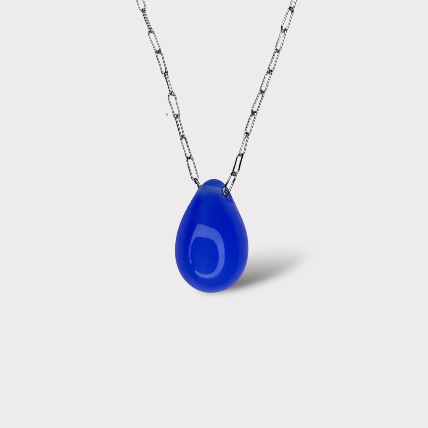 A minimalist necklace featuring an indigo blue teardrop-shaped glass pendant, suspended on a stainless steel chain, displayed on a clean white background.