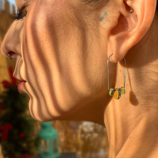 A close-up of a model wearing handcrafted hoop earrings with gradient teardrop glass pendants in shades of green, blue, and amber, styled in natural sunlight for a bold and artistic look.