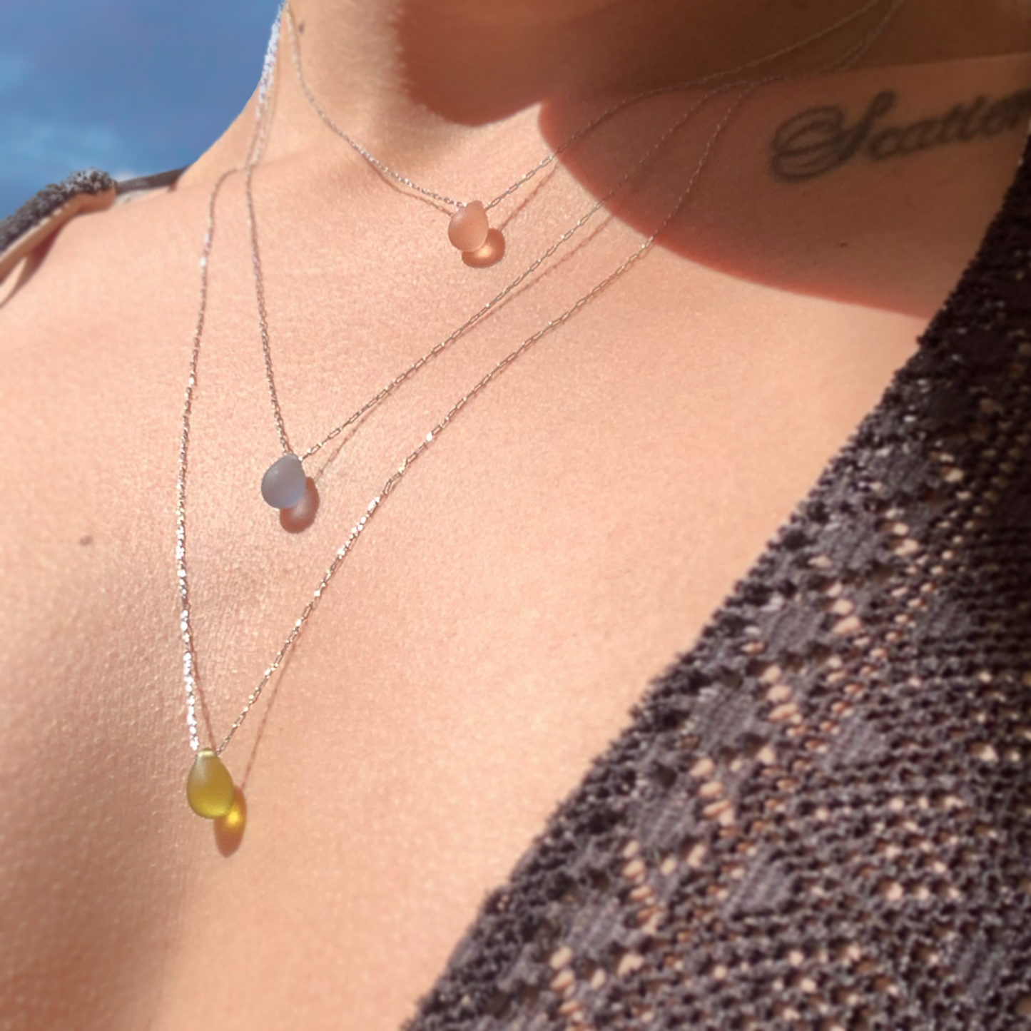 A model wearing a layered set of three teardrop pendant necklaces in matte peach, matte blue, and matte olive glass, suspended on delicate chains, styled in natural sunlight for a radiant look.