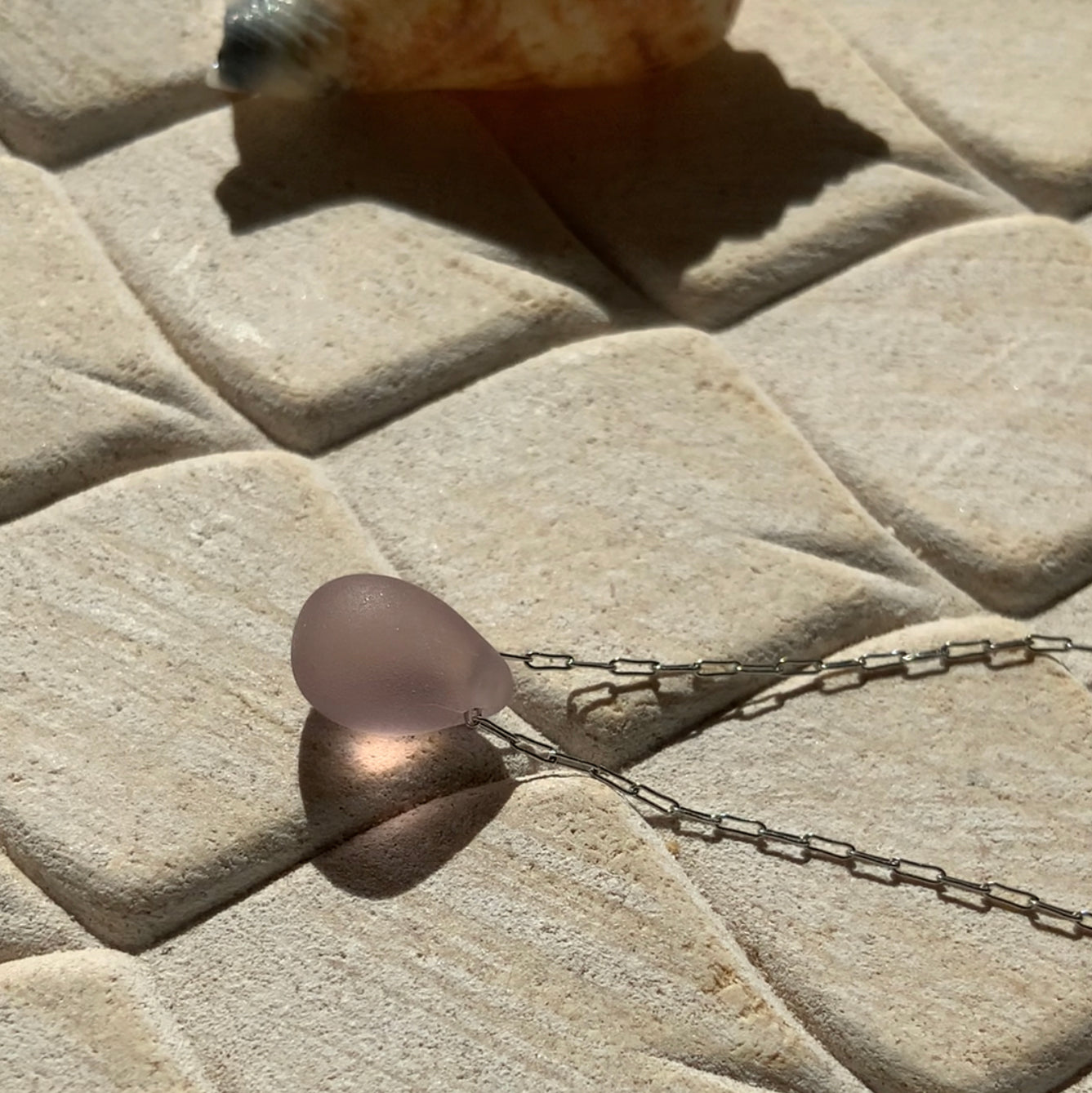 A soft lilac matte teardrop-shaped glass pendant, hanging from a stainless steel chain, displayed on a beige textured background with natural sunlight highlighting its delicate hue.