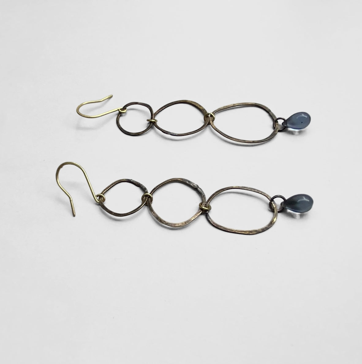 A pair of long brass earrings with a series of oval links, finished with a blue teardrop glass pendant, displayed on a white background for a modern and elegant design.