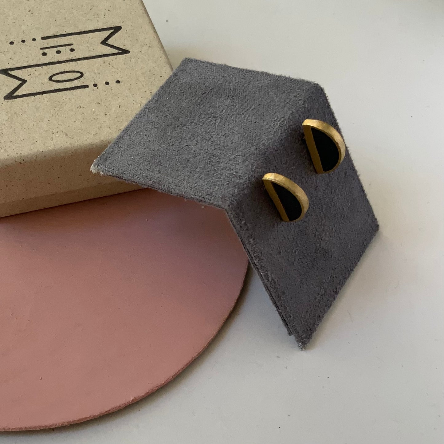 A pair of brass half-cirlce earrings with black accents, elegantly presented on a soft gray stand with branded packaging and a pink accent in the background