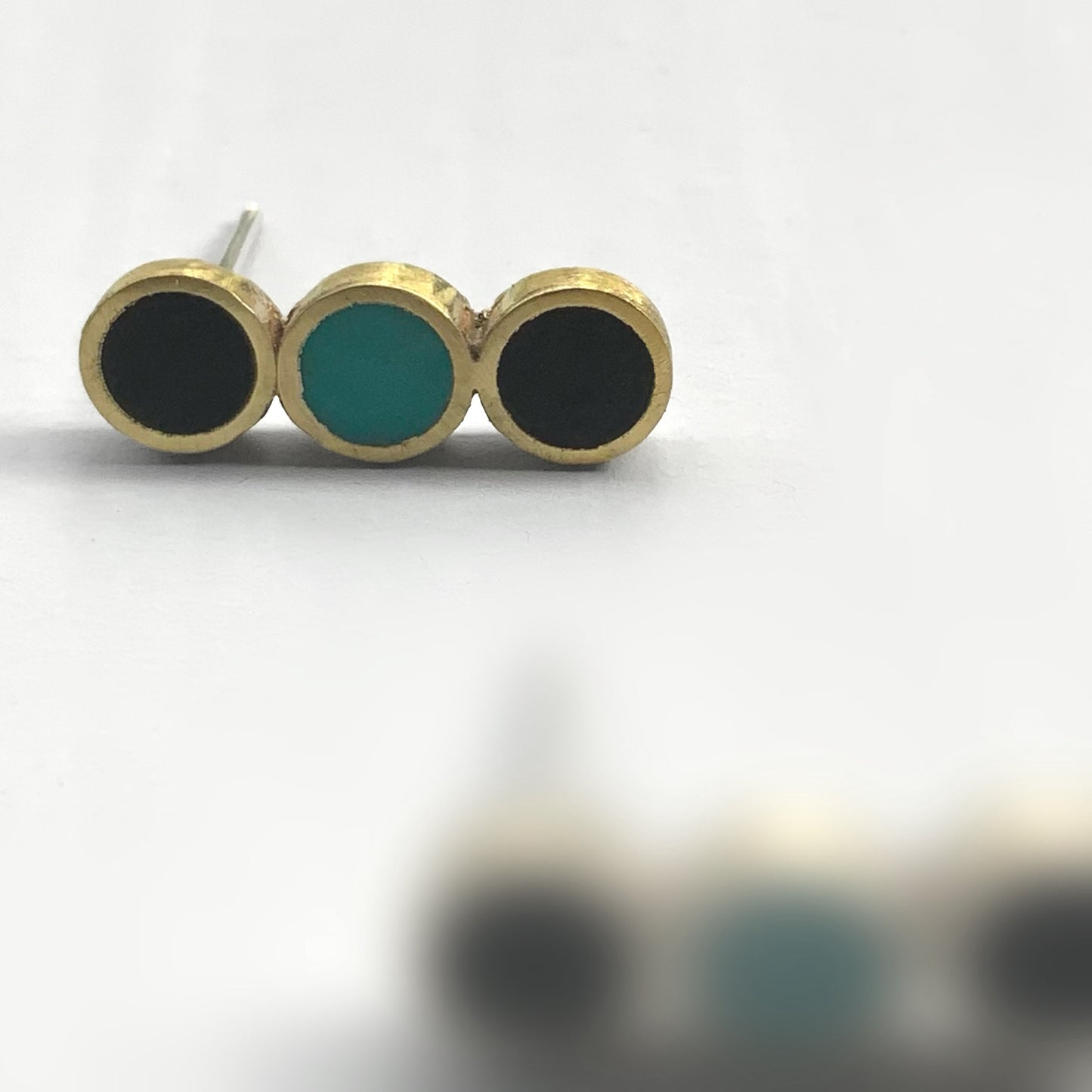 A close-up of brass stud earrings with three circular inlays, two black and one turquoise, arranged in a sleek row, styled on a white background to emphasize their geometric design.