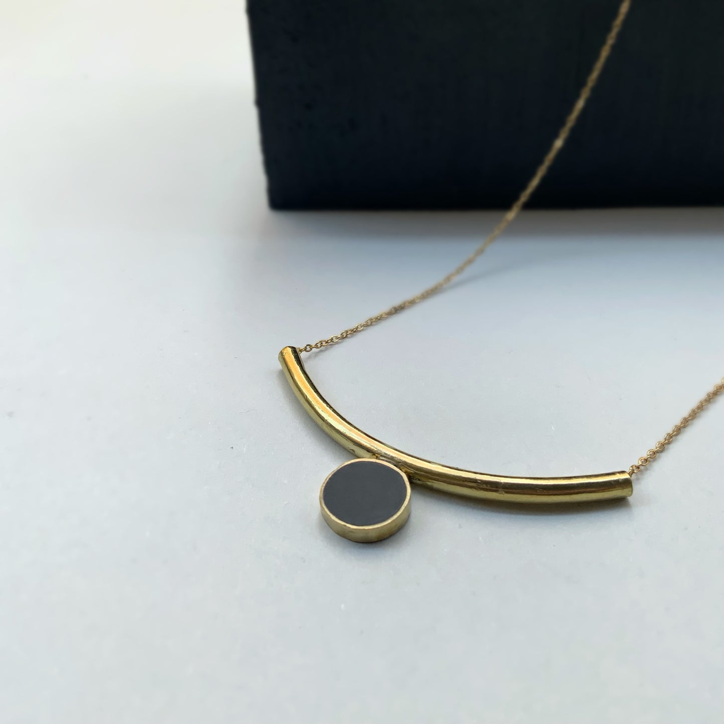A modern brass necklace with a curved arc pendant and a black circular charm, displayed on a soft white background with a dark accent for contrast