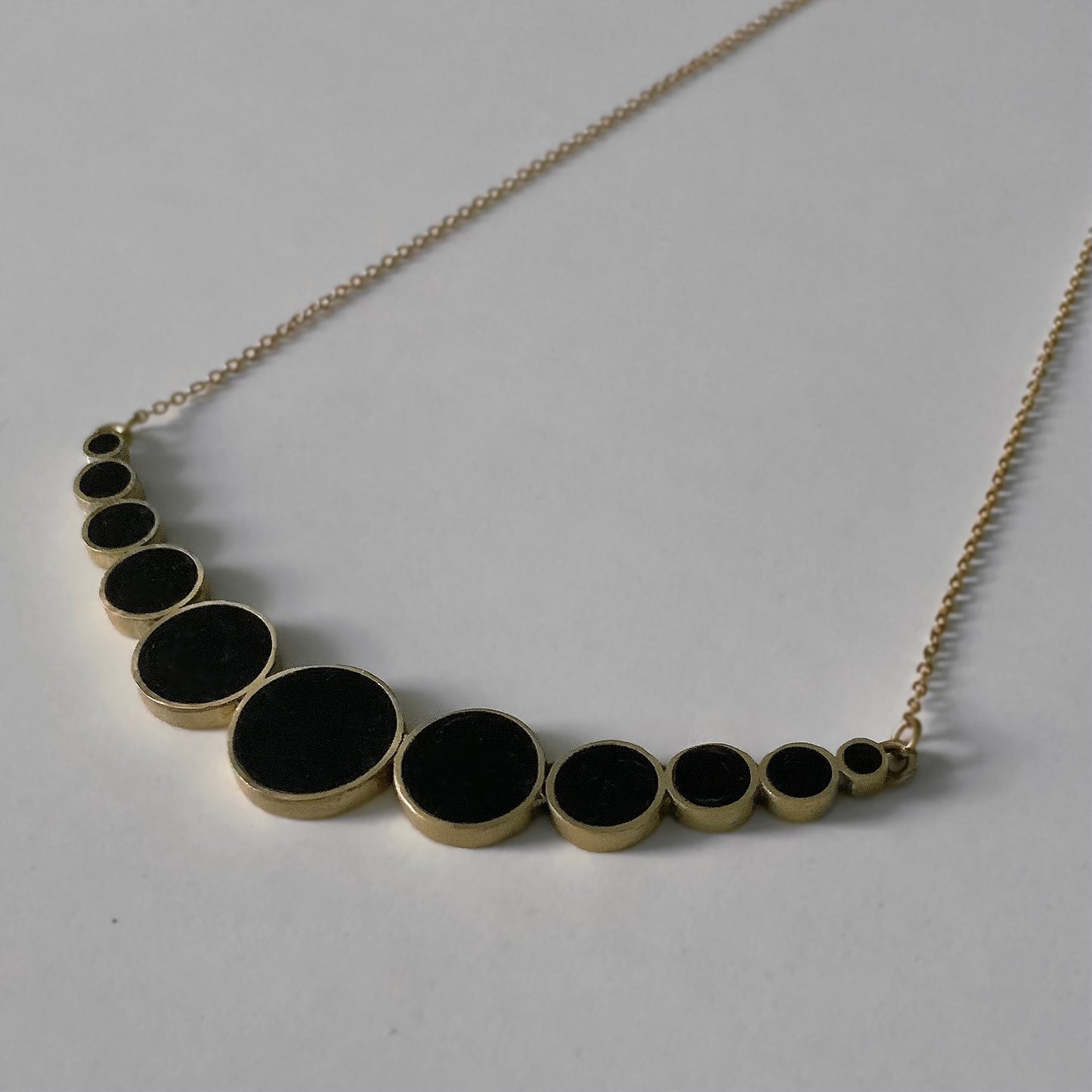 A close-up of a brass necklace with black circular accents in a cascading gradient design, styled on a light gray background to highlight its elegant simplicity