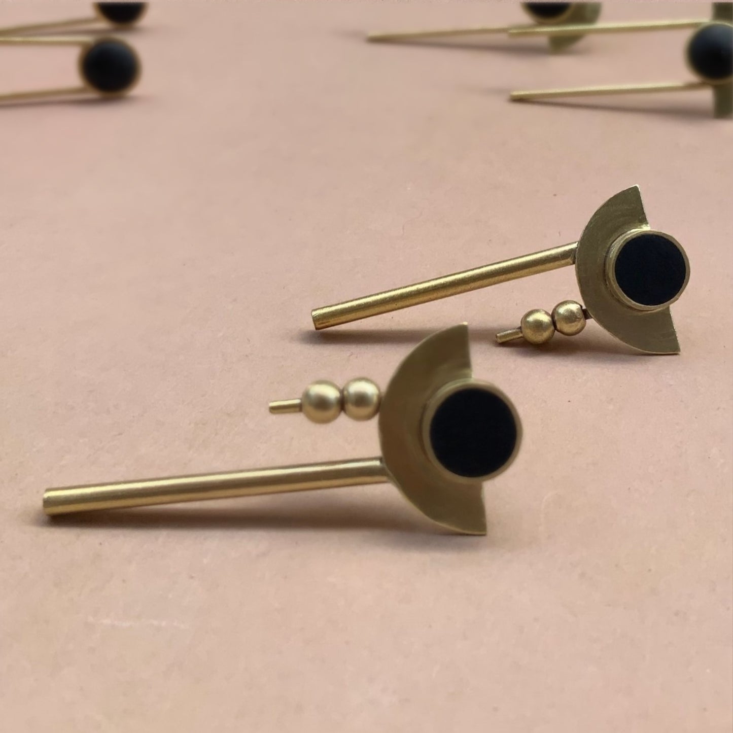 Moon-inspired earrings with black circular centers, brass crescent details, and minimalist rod drops, displayed against a soft pink background with additional earrings blurred in the background for depth
