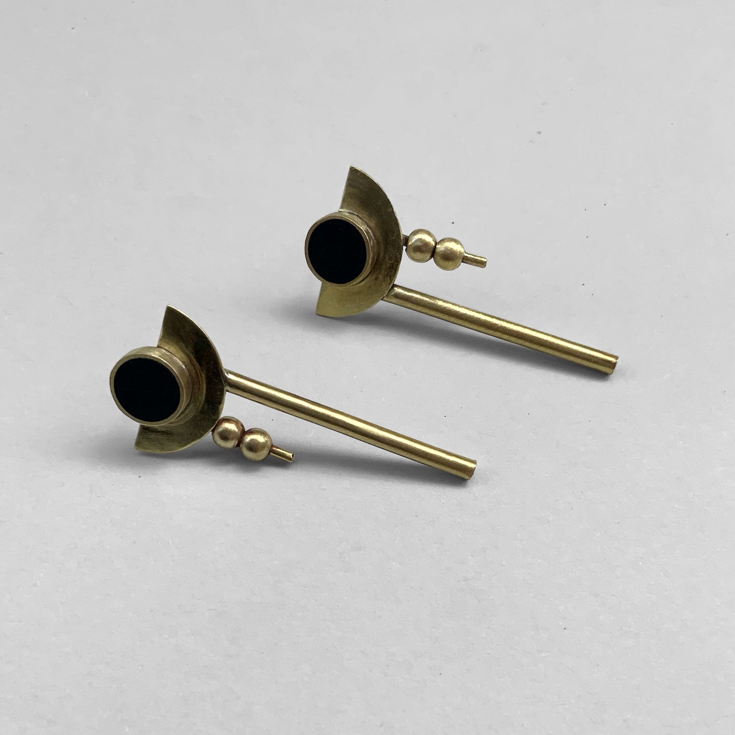 A pair of handcrafted earrings with a black circular centerpiece accented by brass crescent shapes, two small brass beads, and sleek rod drops, arranged on a light gray background