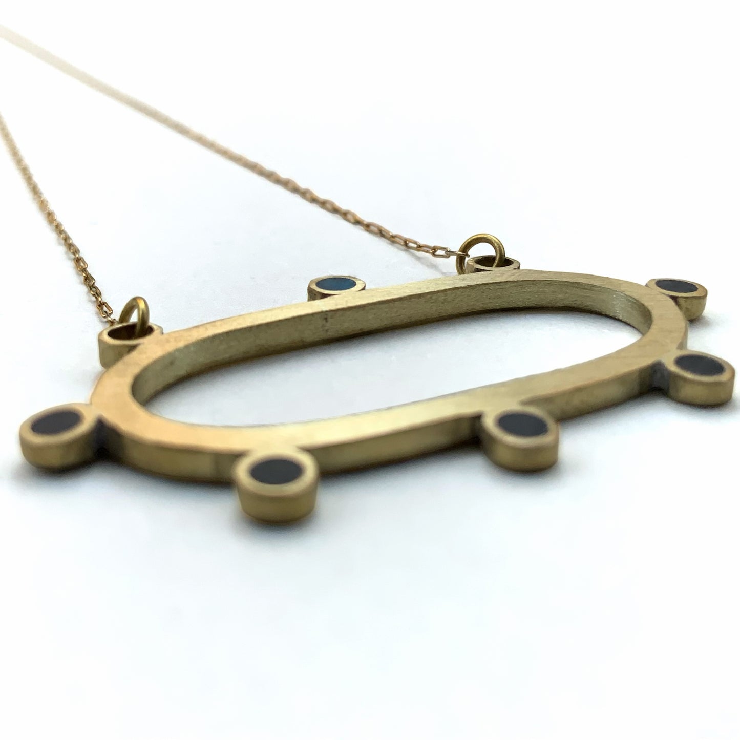 A close-up side view of a brass oval pendant necklace with circular accents, including a subtle blue detail, suspended on a delicate gold chain, photographed against a white background