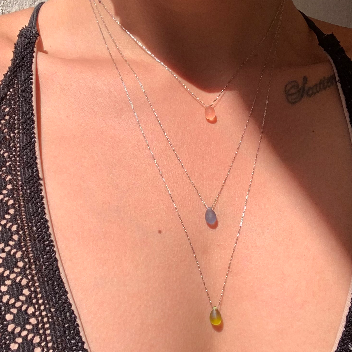A close-up of a model wearing three layered teardrop pendant necklaces in matte peach, matte blue, and matte olive glass, elegantly styled to showcase their minimalist design.