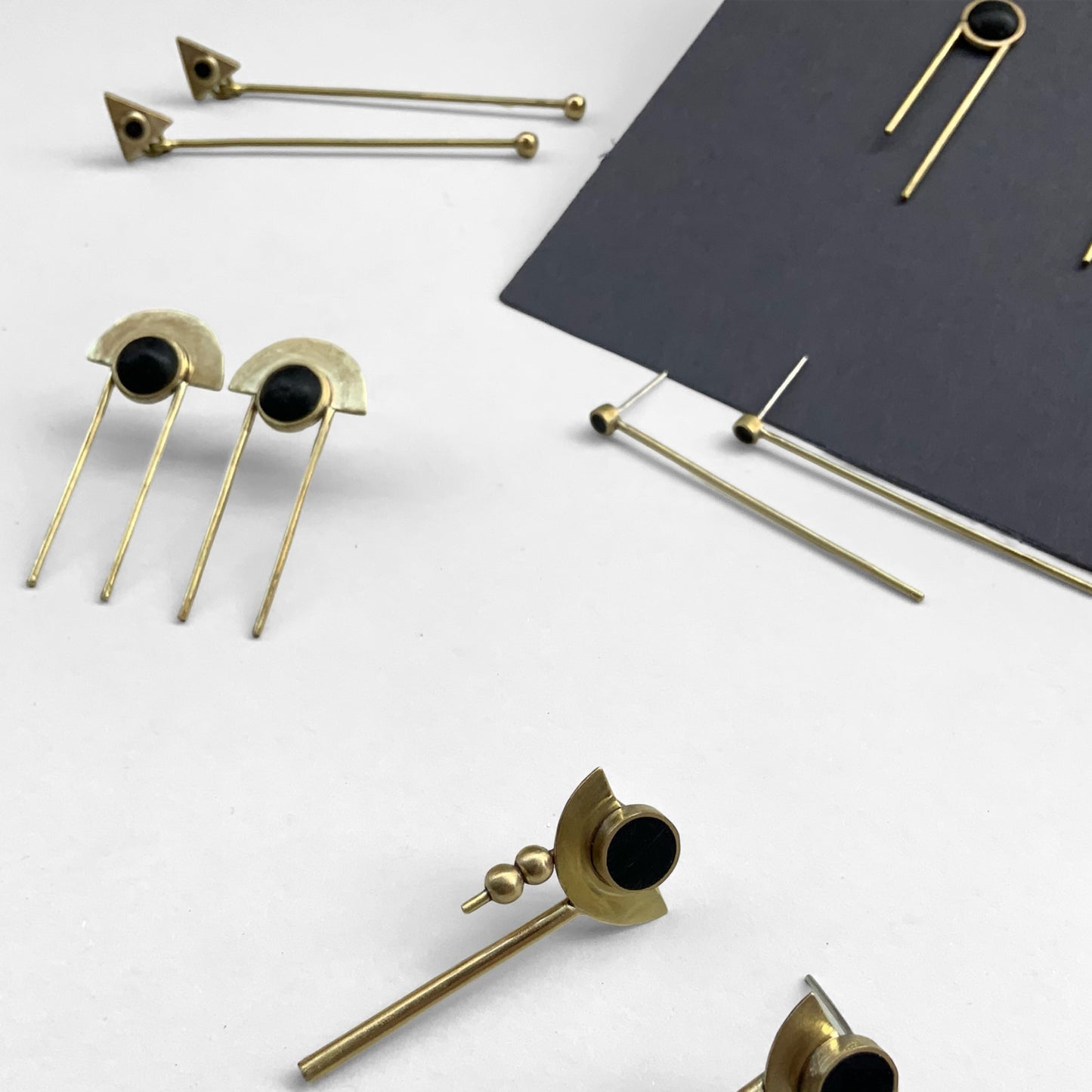 A collection of modern brass earrings, including half-moon designs with black accents and minimalist rods, artistically arranged on a white surface with a dark accent background