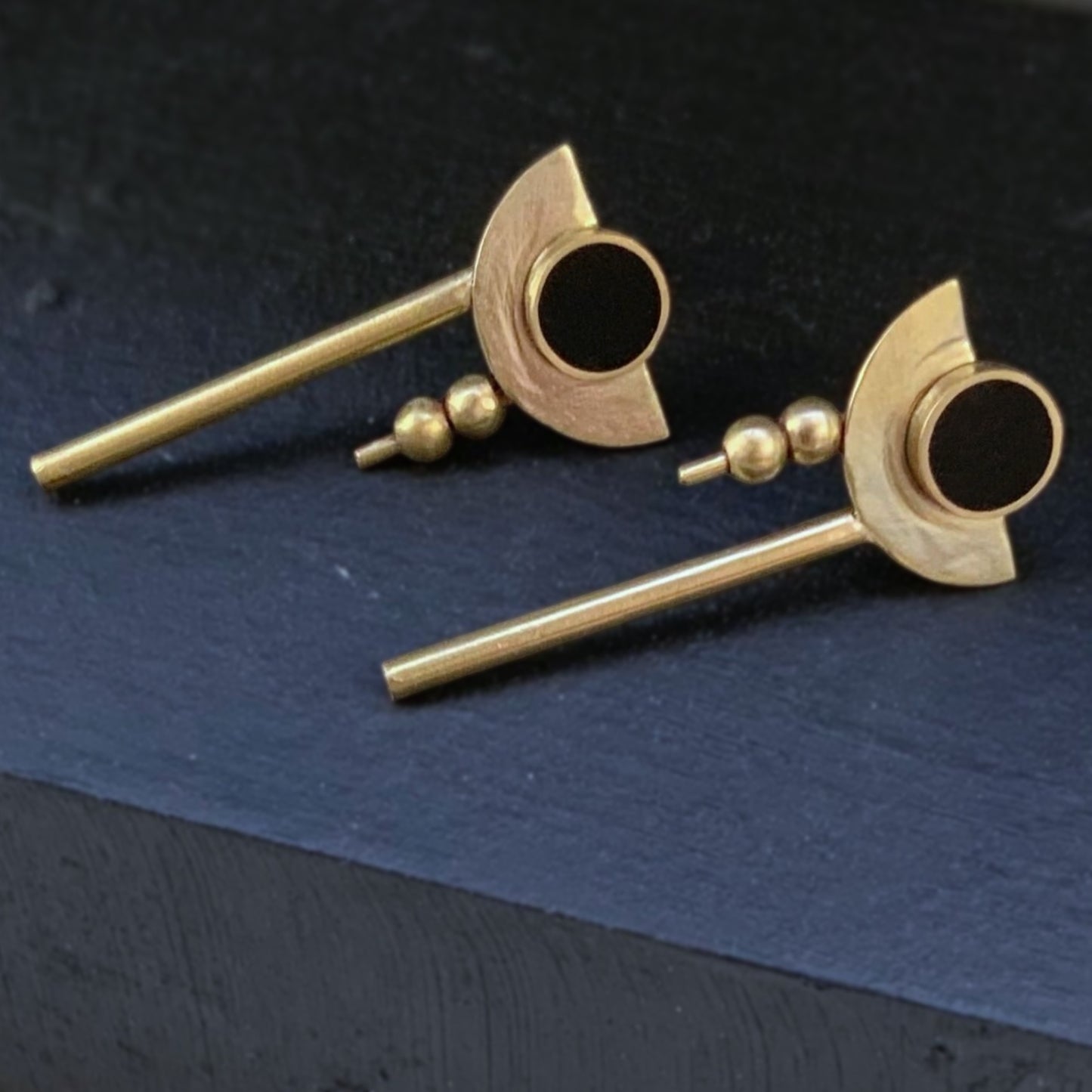Geometric drop earrings with a black circular centerpiece framed by crescent brass details, two small brass beads, and a vertical brass rod, elegantly arranged on a dark textured surface.