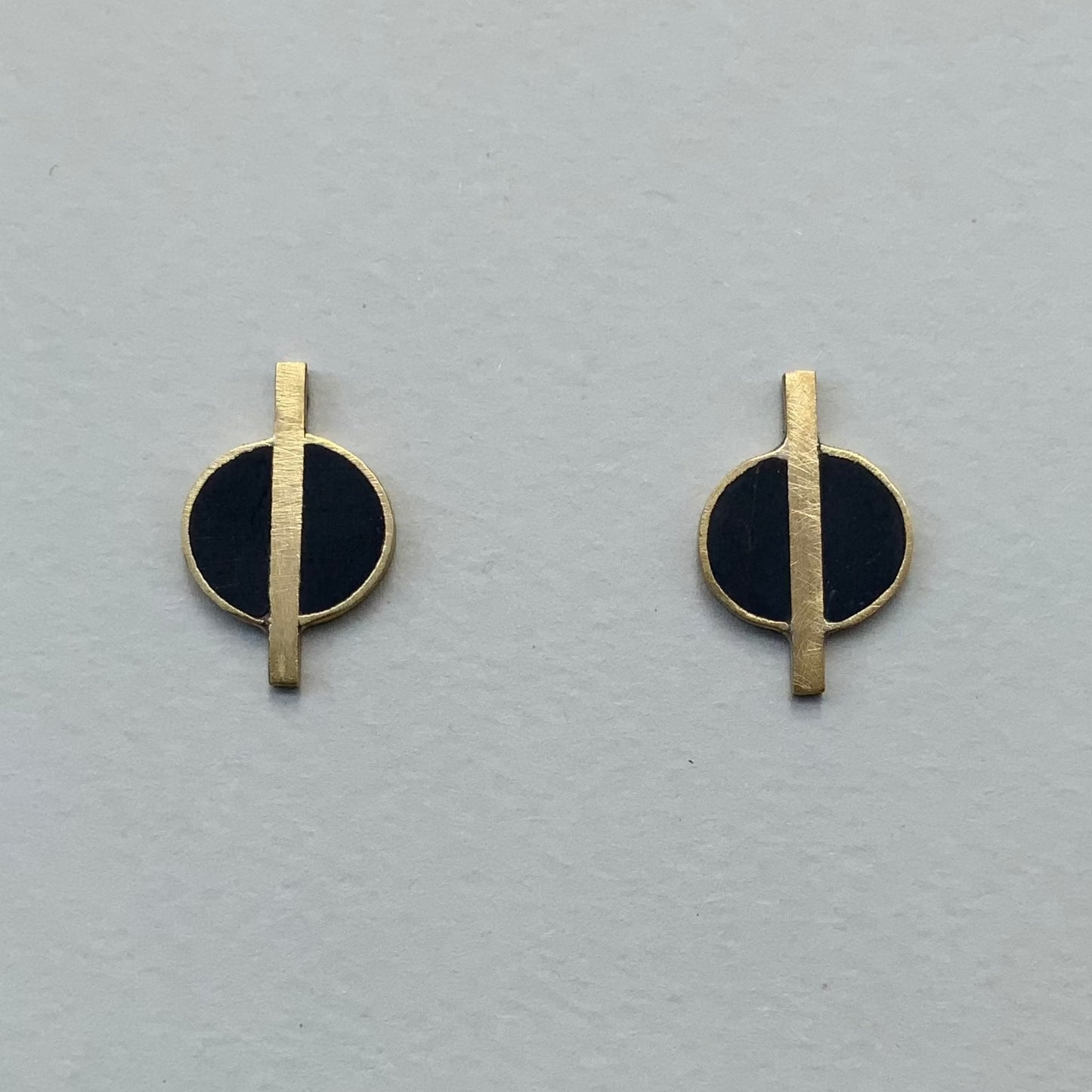 A pair of minimalist brass earrings featuring black circular accents with a vertical brass rod running through the center, displayed on a light gray background