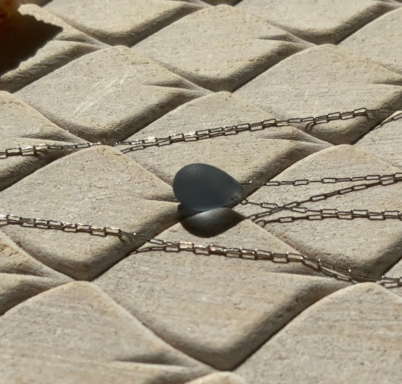 A soft Montana blue matte teardrop-shaped glass pendant, suspended on a stainless steel chain, styled on a beige textured background with natural sunlight highlighting its elegant and minimalist design.