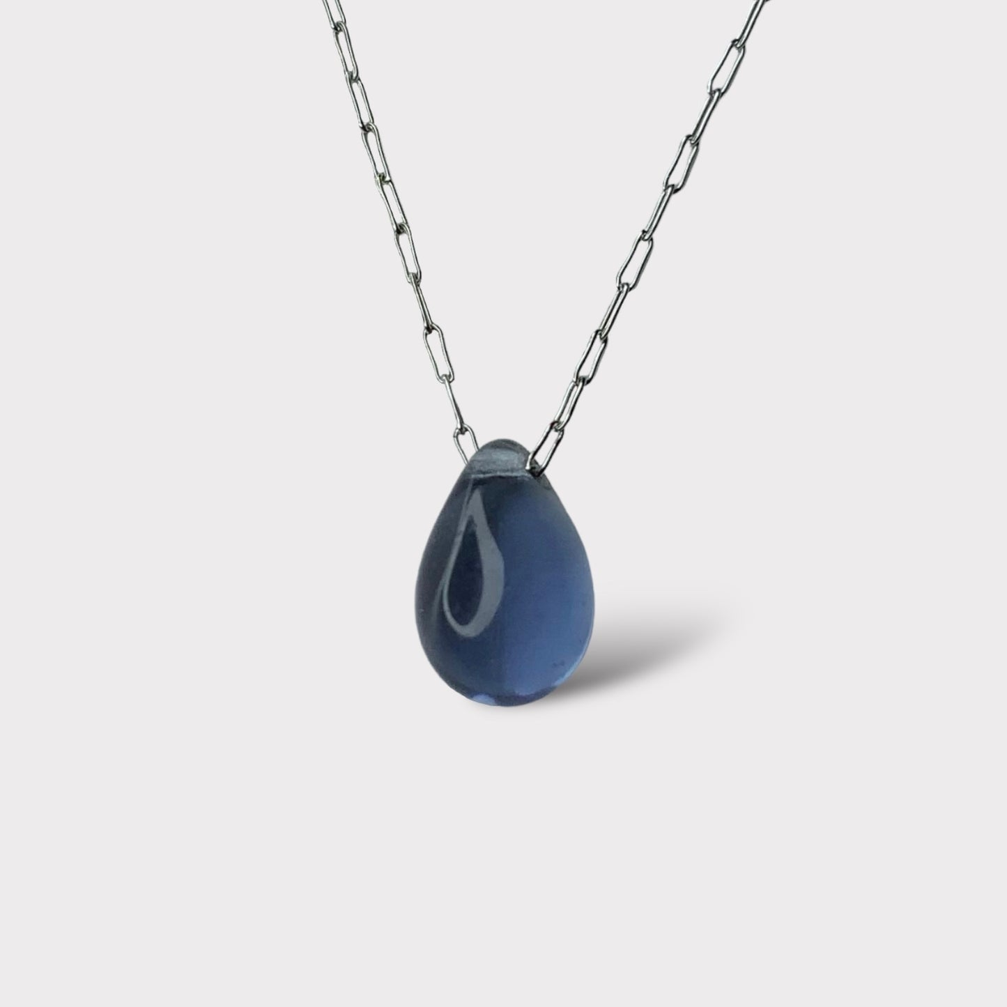 A sleek Montana blue translucent teardrop-shaped glass pendant, hanging from a stainless steel chain, displayed on a white background for a bold and elegant design.