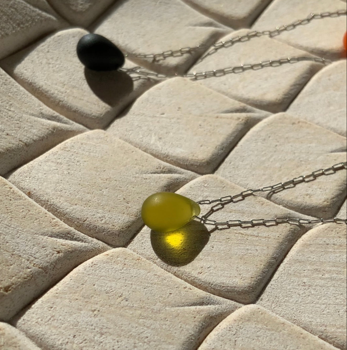 A rich olive matte teardrop-shaped glass pendant, suspended on a stainless steel chain, styled on a beige textured background with natural sunlight creating a warm glow.
