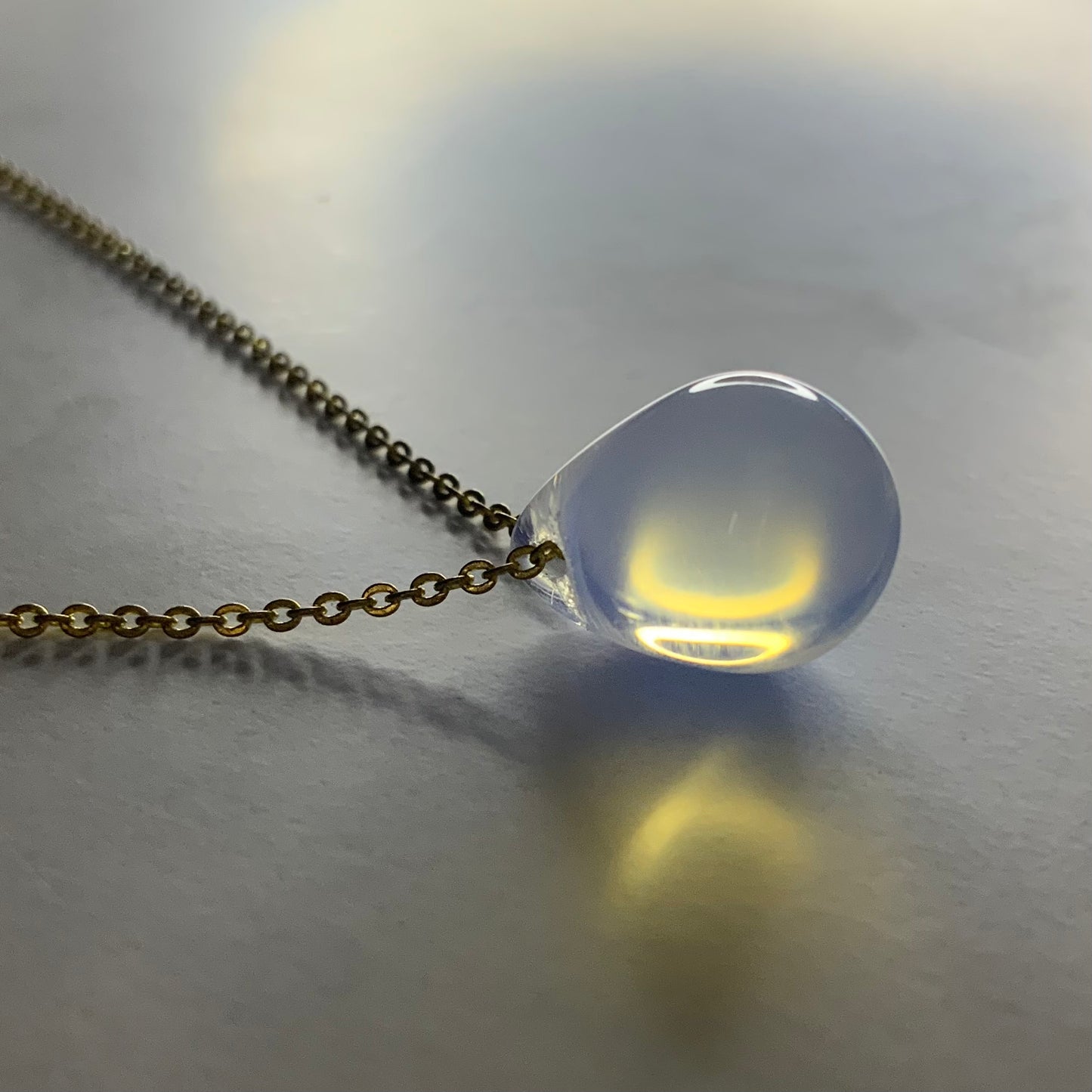 A handcrafted necklace with an opal teardrop glass pendant, suspended on a brass chain, displayed on a softly lit surface for a vibrant and artistic design.