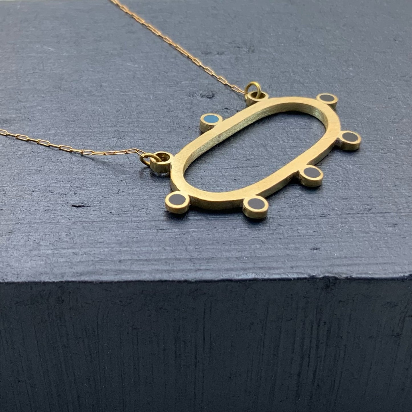 A handcrafted brass necklace featuring an elongated oval pendant with circular accents and a delicate blue detail, suspended on a fine golden chain, styled against a dark slate background