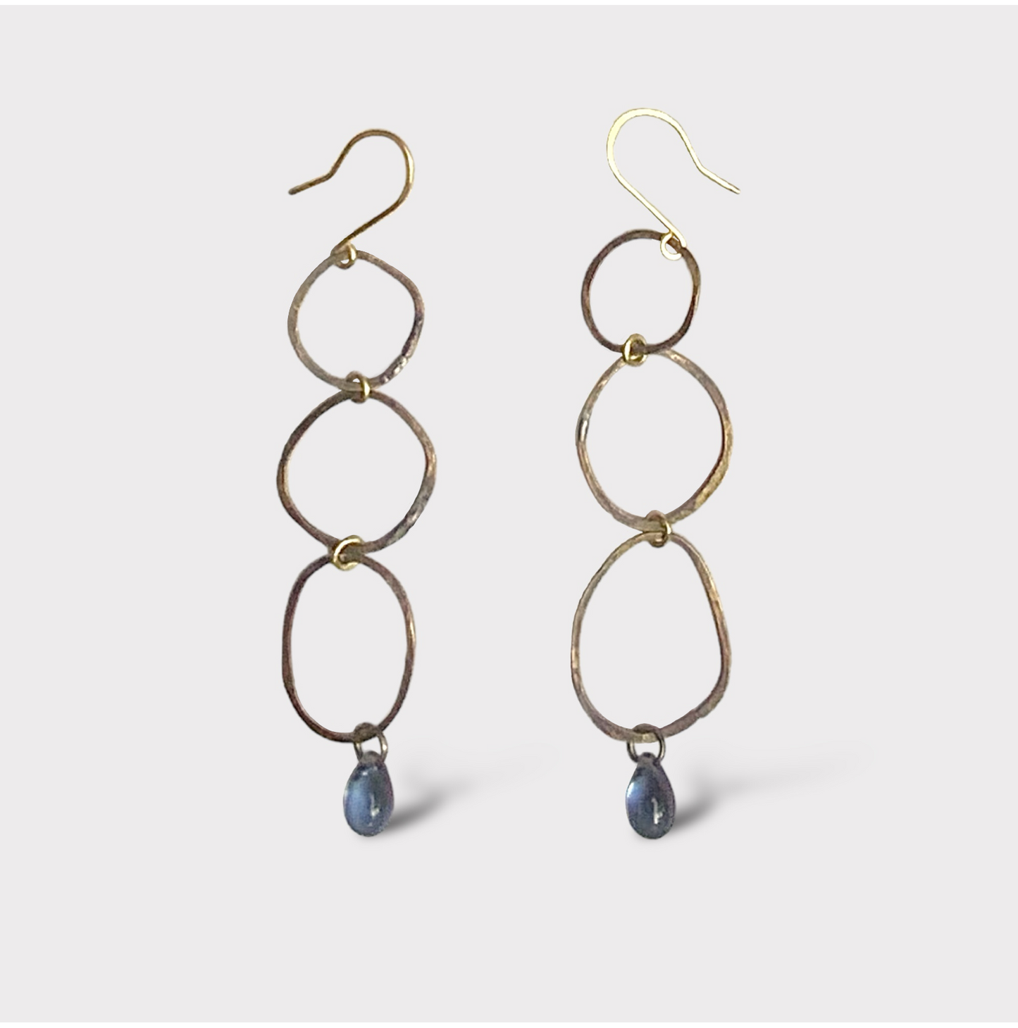 A pair of handcrafted earrings featuring interconnected oval brass links with a blue teardrop glass pendant at the bottom, suspended on gold hooks, styled on a white background for a minimalist look.